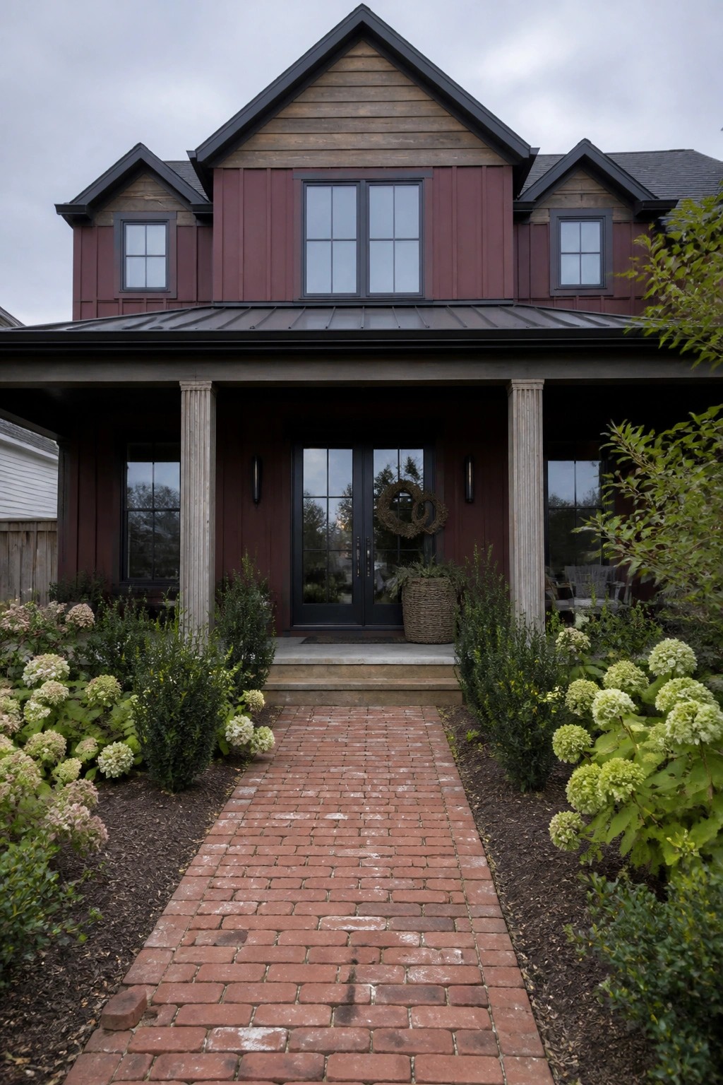 A two-story house with smooth burgundy fiber cement siding, black-framed windows and doors, a covered porch with columns, and a red brick walkway.