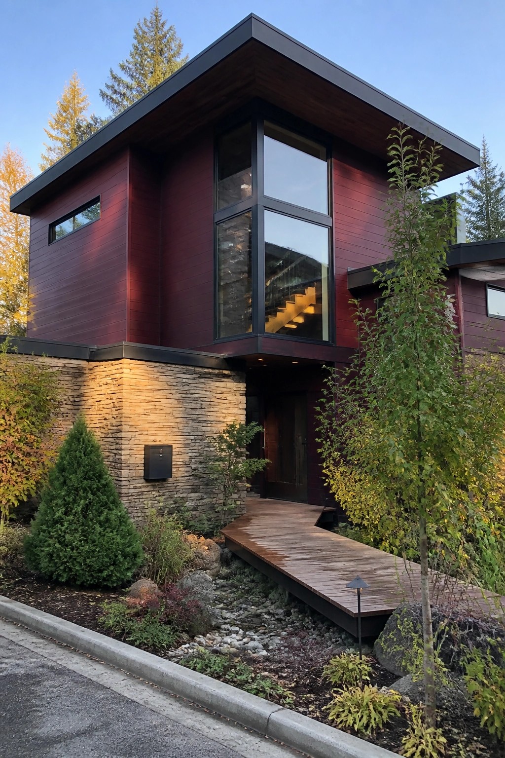 A modern two-story house with deep burgundy fiber cement siding on the upper level, a stone foundation, large black-framed windows, and a wooden walkway leading to the front door.