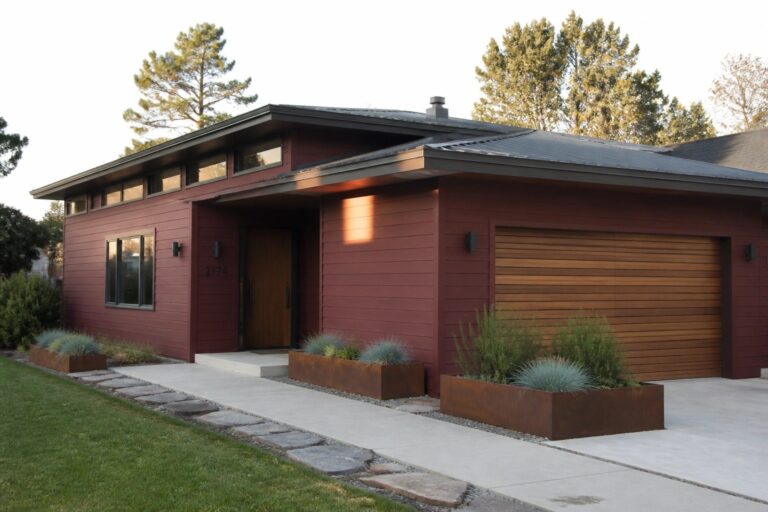 19 Modern Maroon House Exterior Ideas Using Smooth Fiber Cement Siding A modern single-story house with maroon horizontal fiber cement siding, a dark metal roof, a wooden front door, and a wooden garage door, with concrete walkway and metal planters holding shrubs.