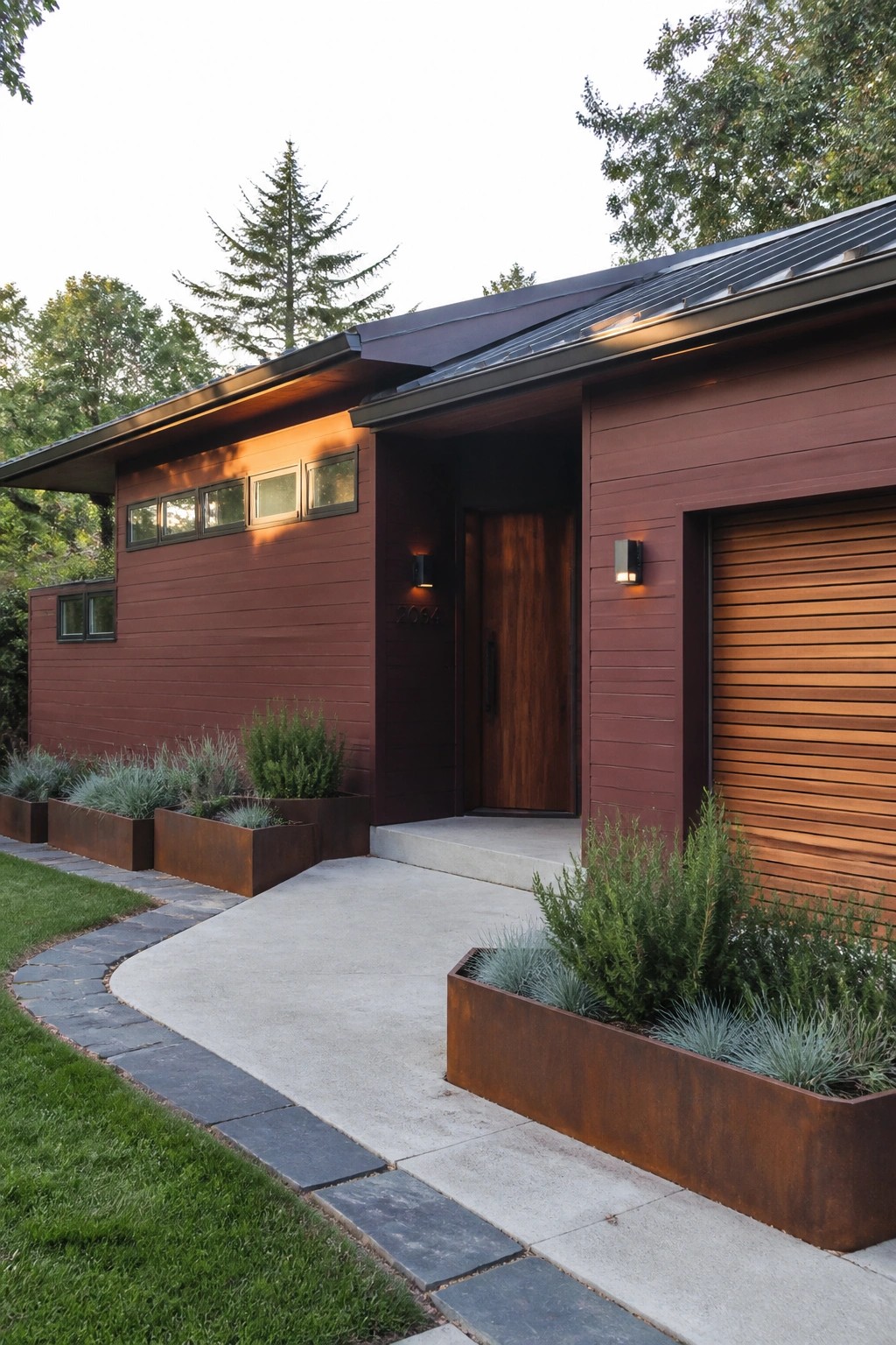 A modern single-story house with maroon horizontal fiber cement siding, a dark metal roof, a wooden front door, and a wooden garage door, with concrete walkway and metal planters holding shrubs.