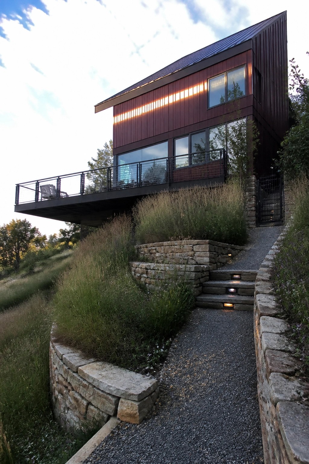 Modern red house with balcony above stone steps and gravel path on hillside