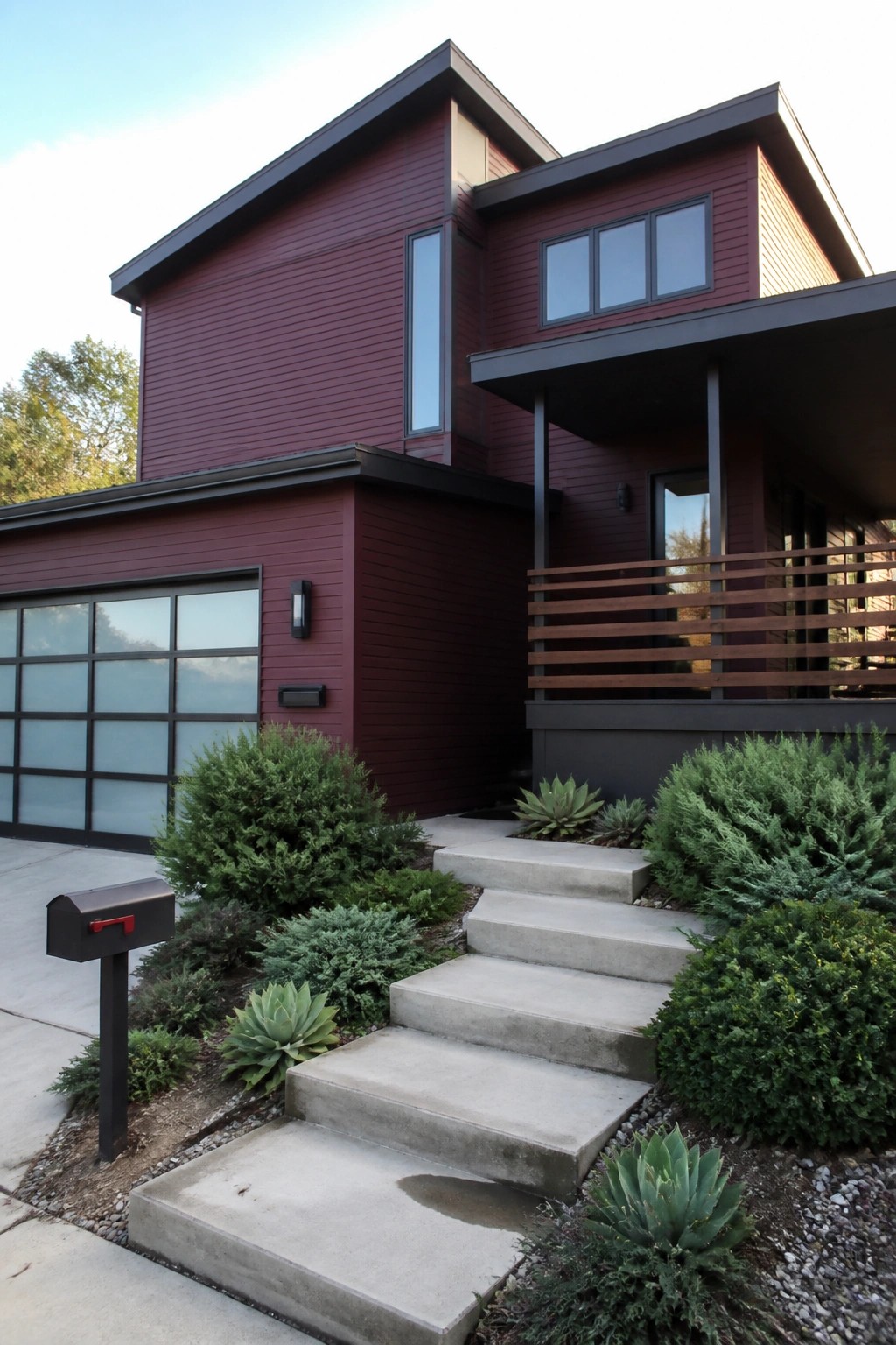A two-story modern house with maroon fiber cement siding, a concrete staircase, and a dark garage door.