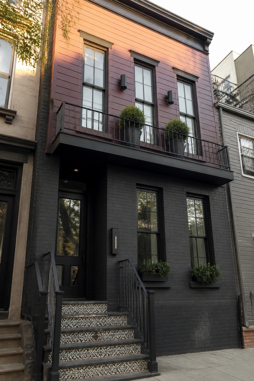 A narrow modern house with smooth maroon fiber cement siding on the upper facade and black brick below, shown with a second floor balcony and black window frames.