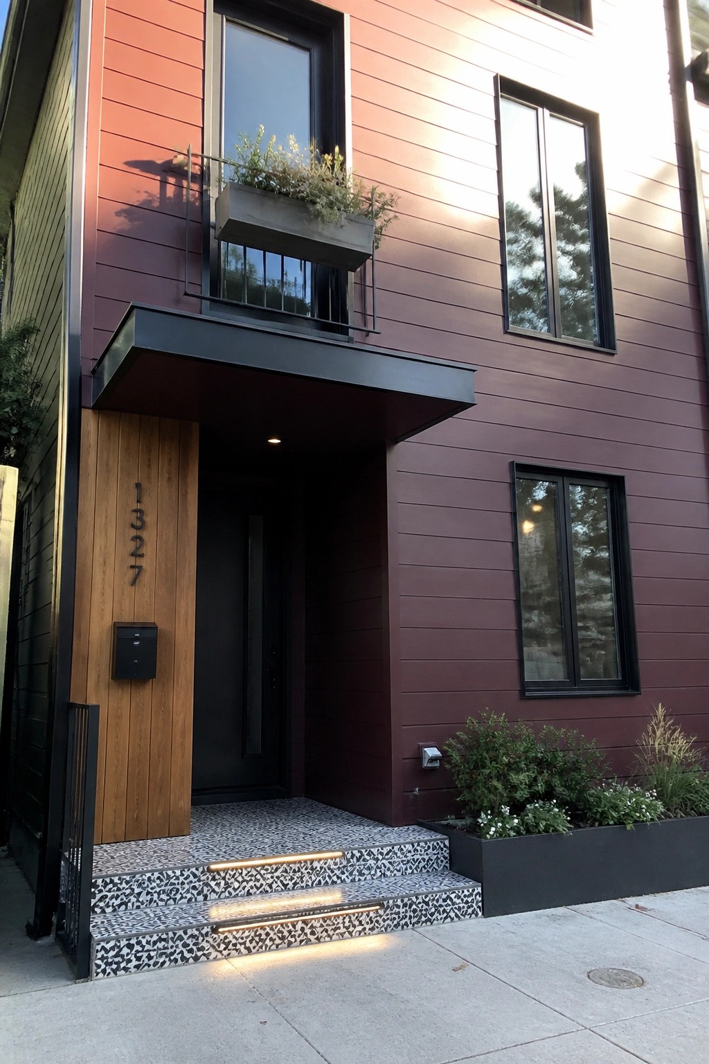 Modern home entrance with red siding, black door, mosaic steps, and balcony planter.