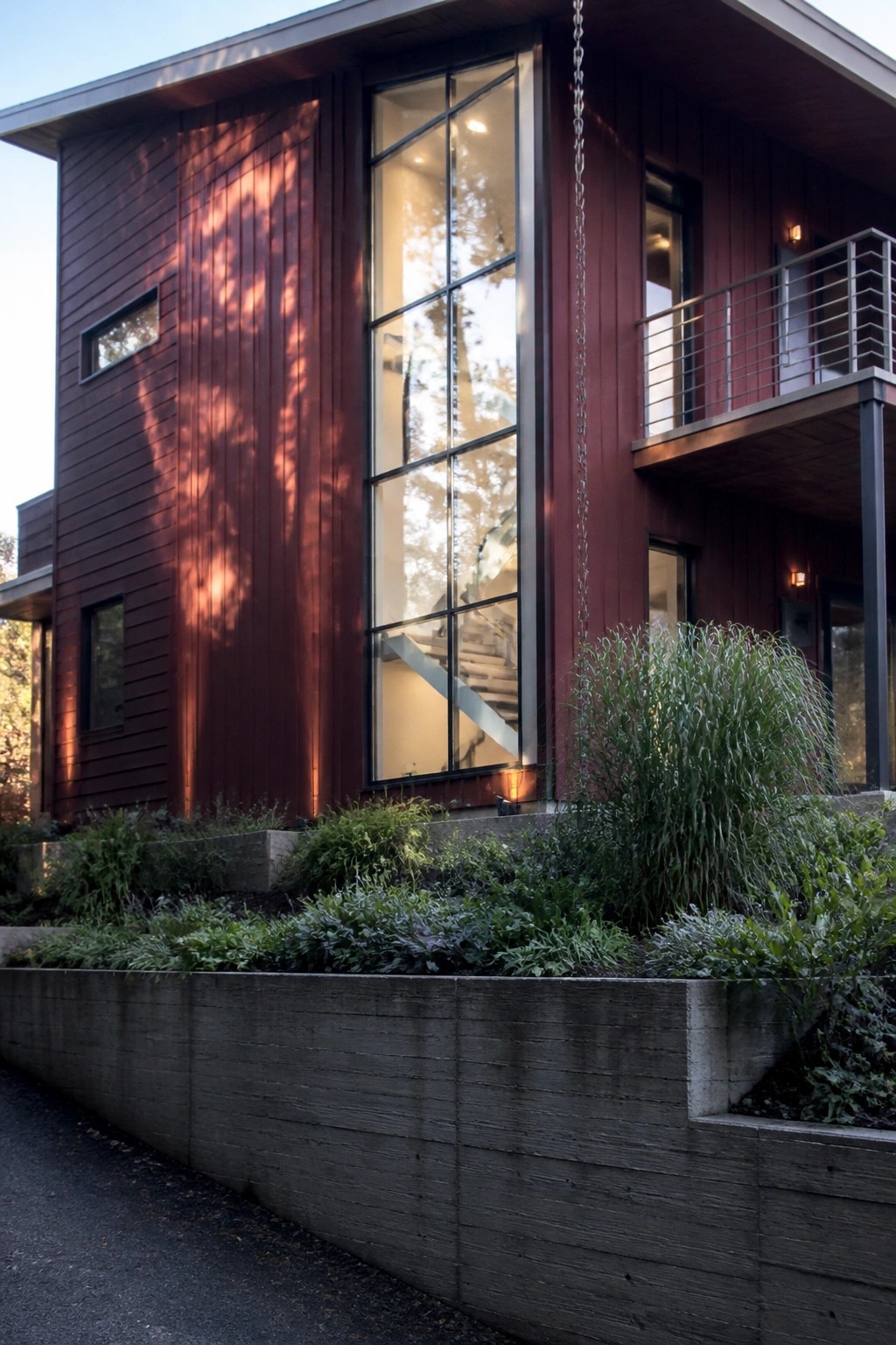 A modern two-story house clad in vertical maroon fiber cement siding with tall black-framed windows and a cantilevered balcony.