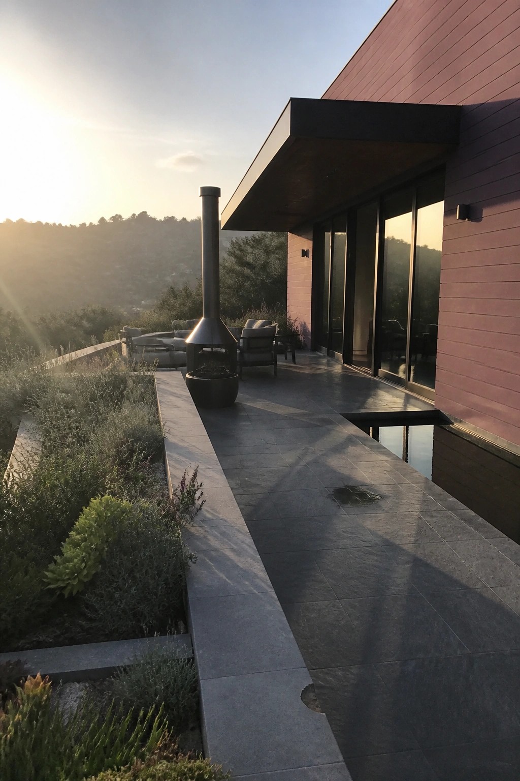 Modern house with stone patio, outdoor fireplace, garden, and hill views at sunset