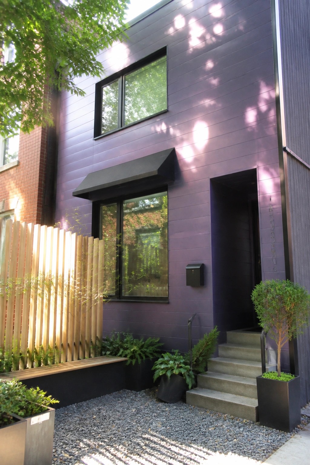 A two-story house with deep purple horizontal siding, black-framed windows of varying sizes, a black entry door with concrete steps, and gravel ground cover with potted plants.