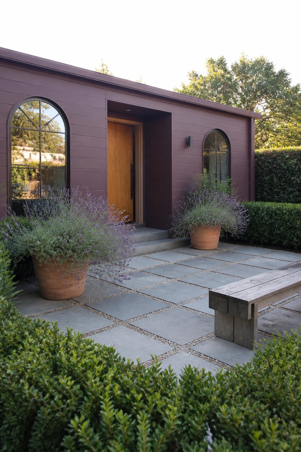 Maroon house with arched windows, wooden door, lavender pots on stone patio with bench.