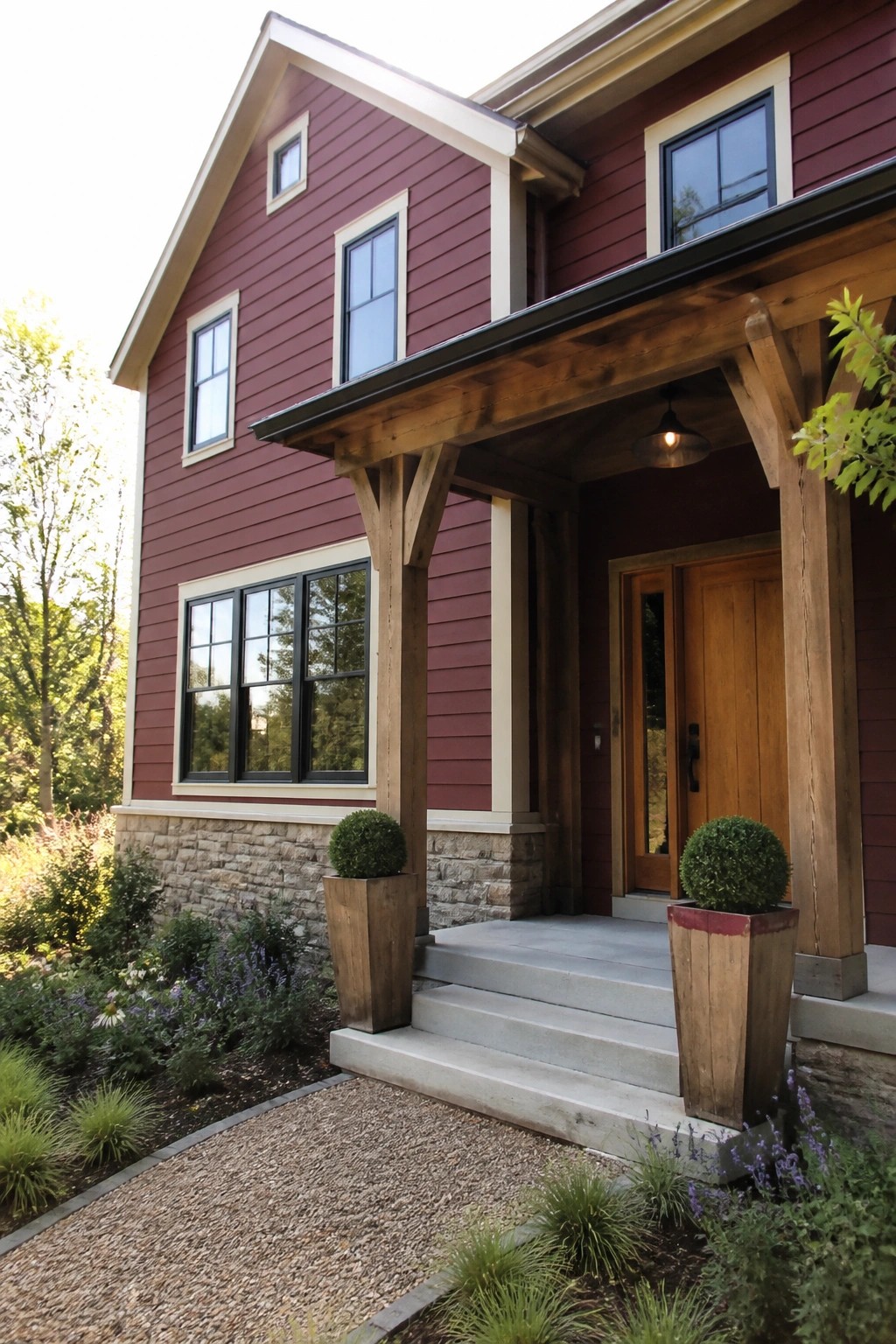 A two-story house exterior with deep burgundy horizontal lap siding, a wooden front porch supported by timber beams, a stone foundation, and potted shrubs flanking the concrete steps.