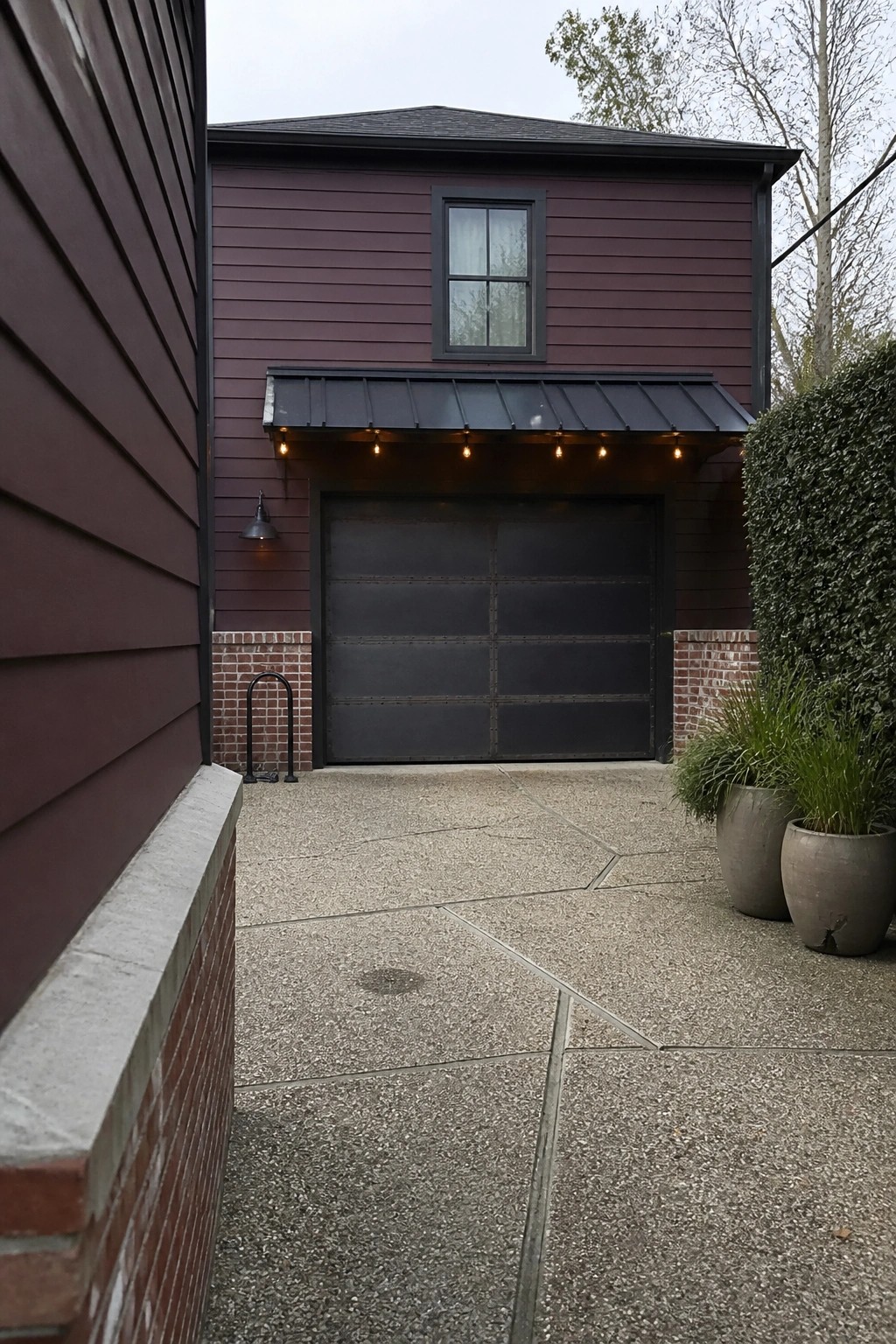 A two-story house with burgundy horizontal lap siding, a dark metal garage door under a black awning with string lights, and brick accents at the base.