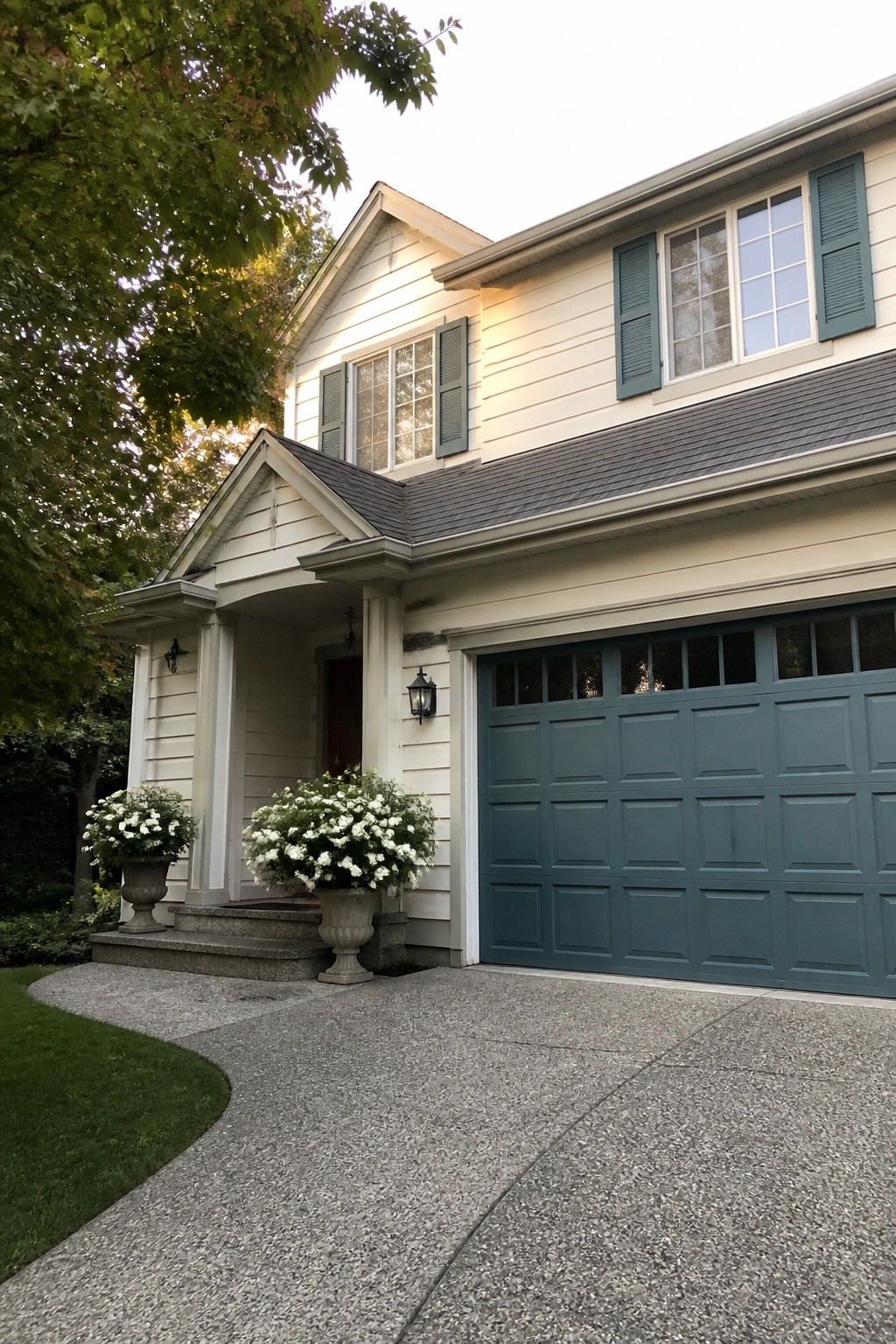 Beige two-story house with green garage door, shutters, and white flower urns.