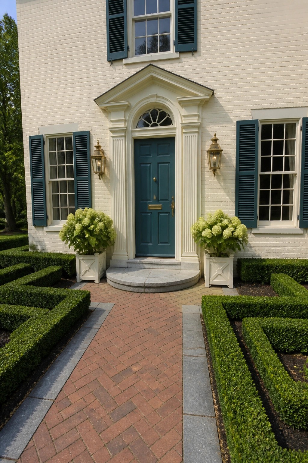 Front view of a white brick house with a teal door centered under a cream pediment, teal shutters on the windows, and two white planters with hydrangeas beside the steps.