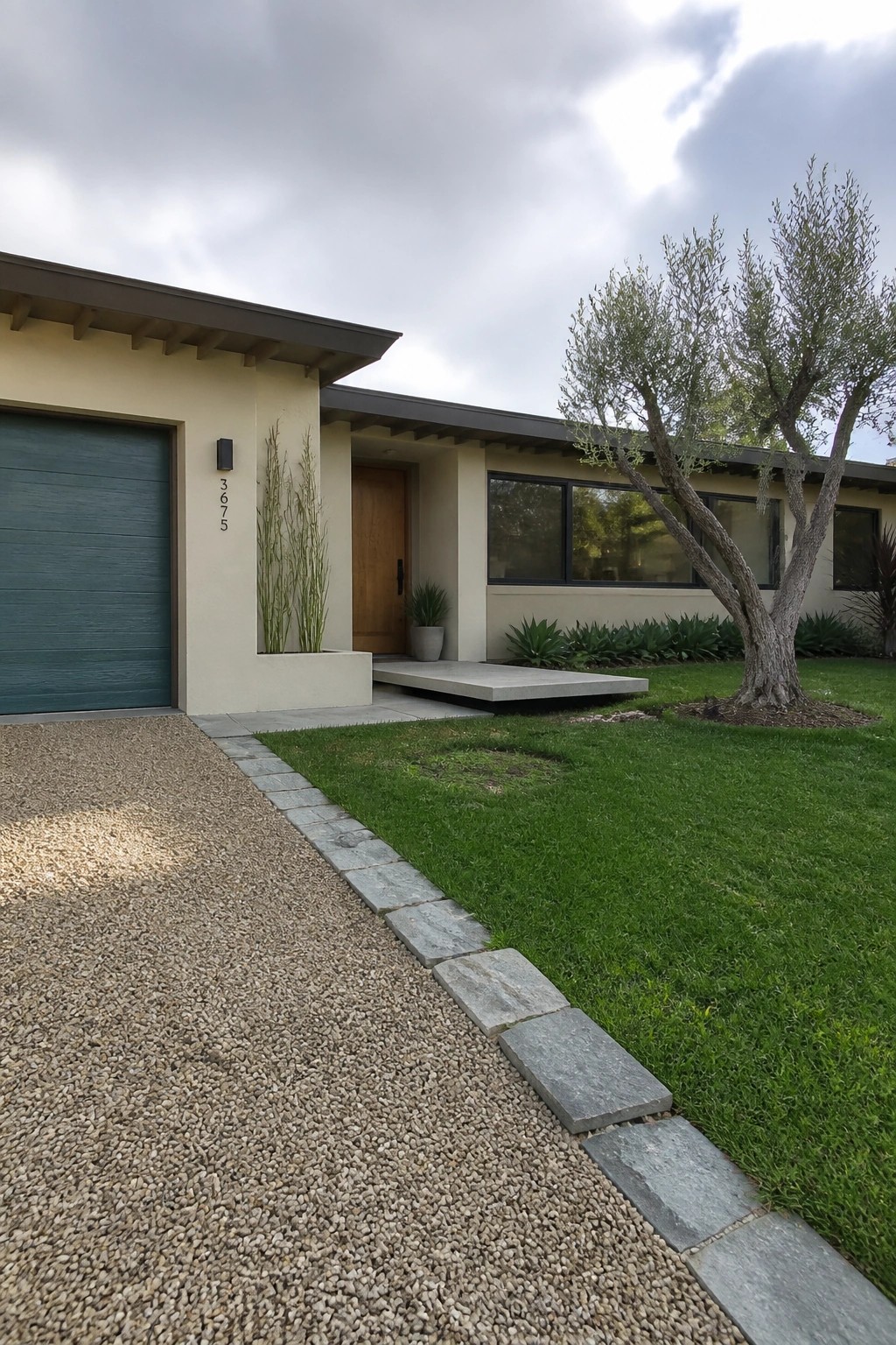 Modern beige house with teal garage, wooden door, gravel driveway, and green lawn.