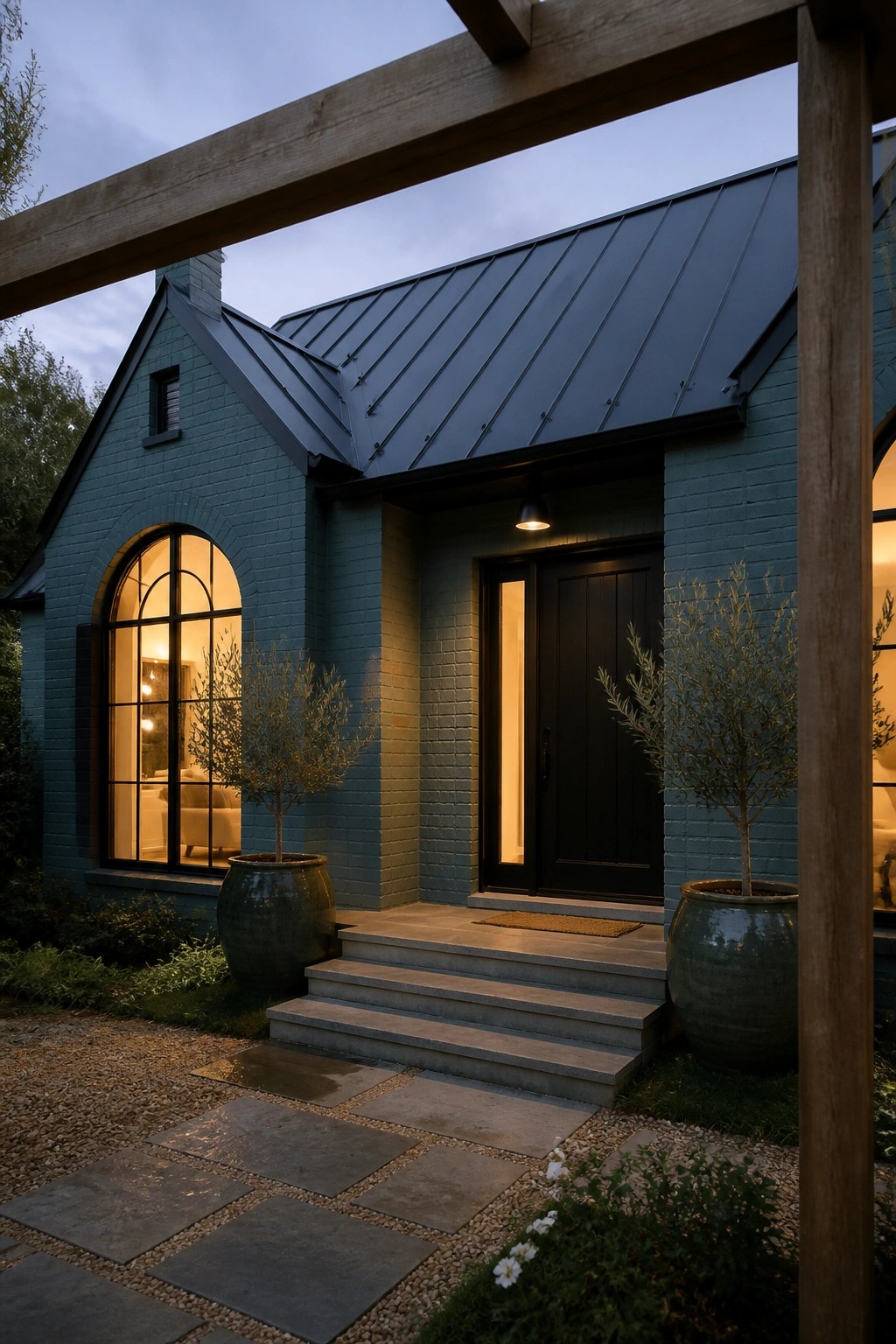 A teal brick house exterior with a black metal roof, black front door, arched window, and concrete steps flanked by large potted plants.