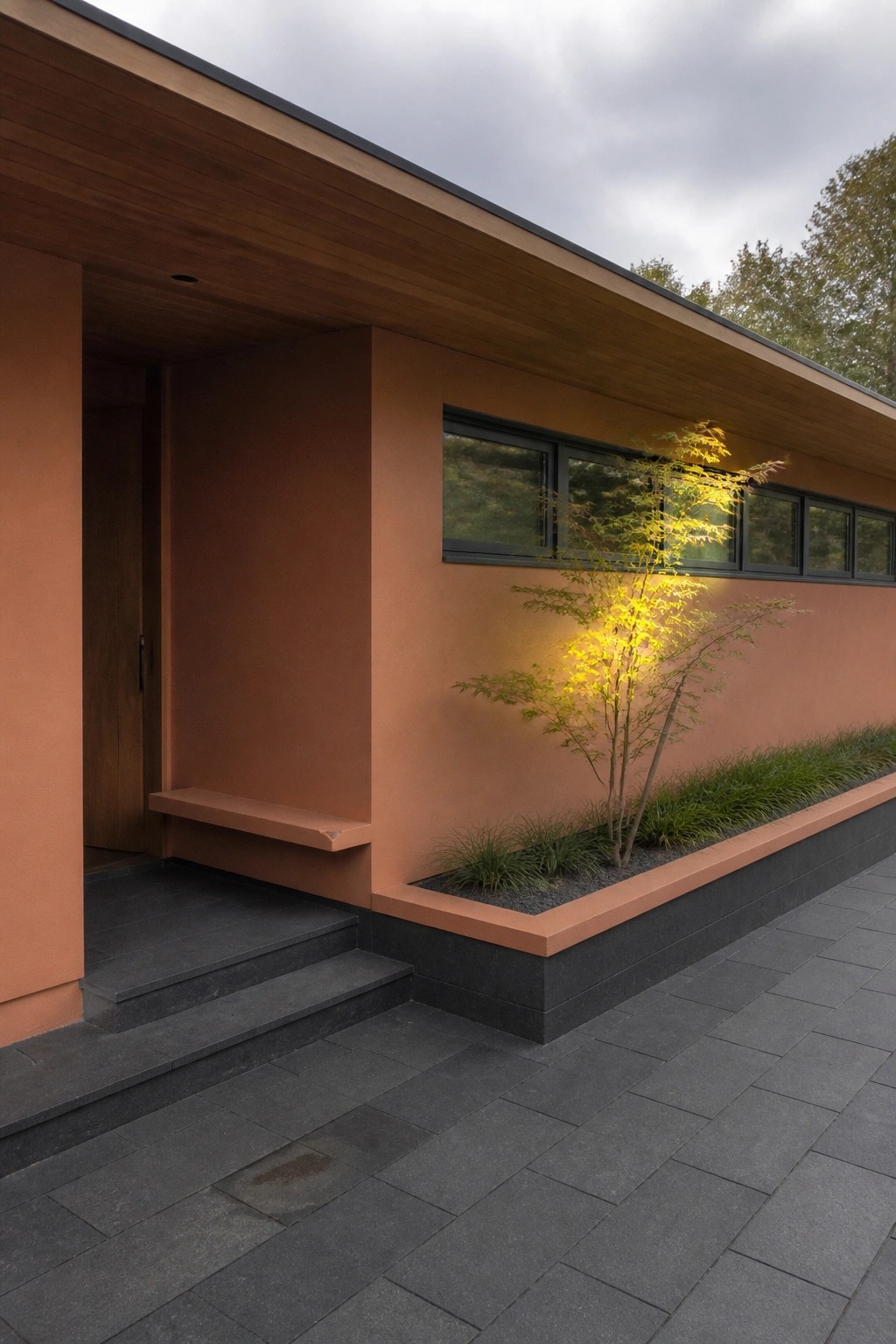 Exterior view of a single-story ranch house with orange stucco walls, a long wooden roof overhang, horizontal windows, a built-in bench, and an integrated planter bed.