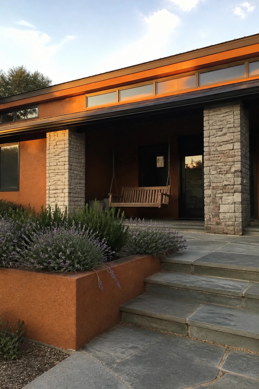 A single-story ranch house exterior with orange stucco walls, a flat roofline, stone pillars supporting a covered porch with a wooden swing, and stone steps in the foreground.