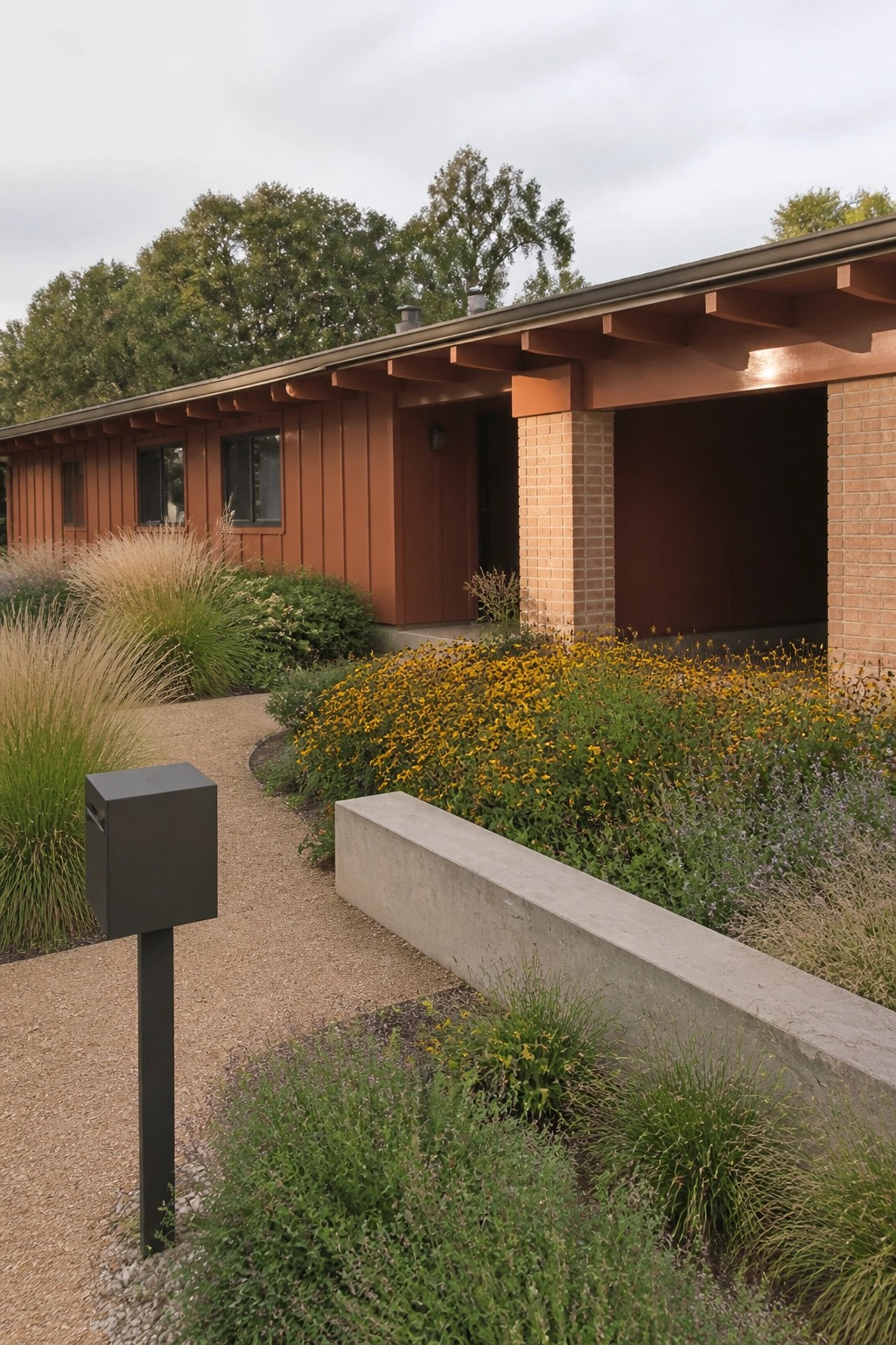 A single-story ranch house with orange-brown siding, brick supports, and a low roofline sits next to a gravel path bordered by yellow flowers and tall grasses.