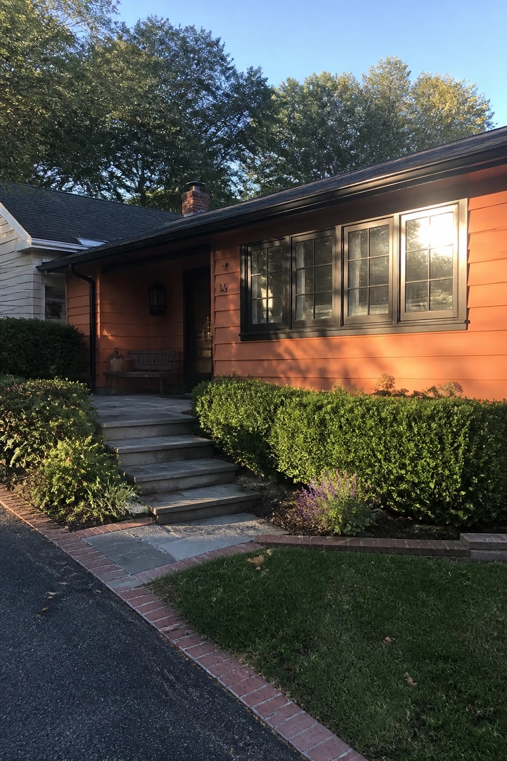 Orange house with black windows, stone steps, green shrubs, and brick edging in sunlight