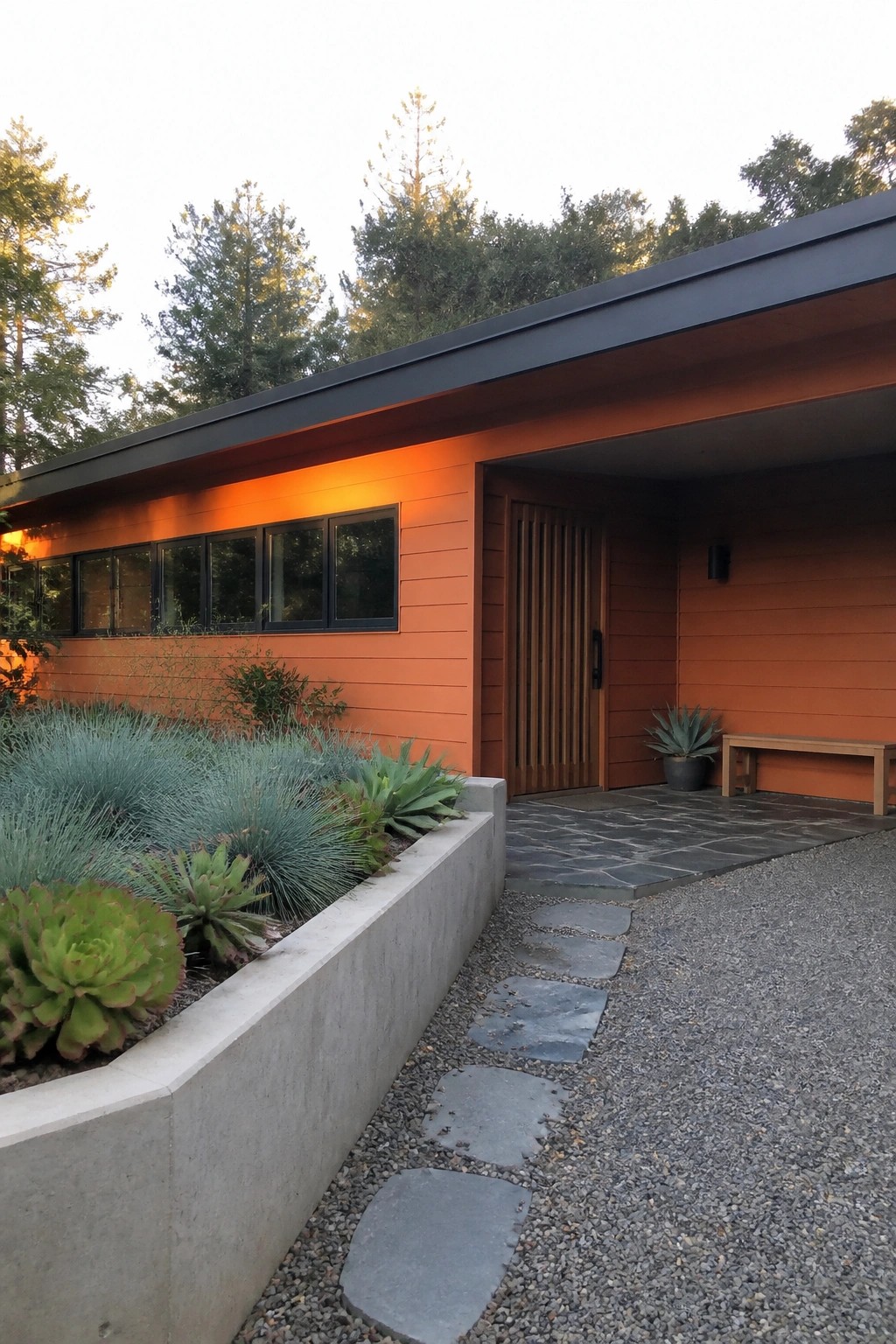 A single-story ranch house exterior with orange horizontal siding, a low black roof overhang, a row of black-framed windows, a vertical wood slat door, and a concrete planter with succulents beside a stone path.