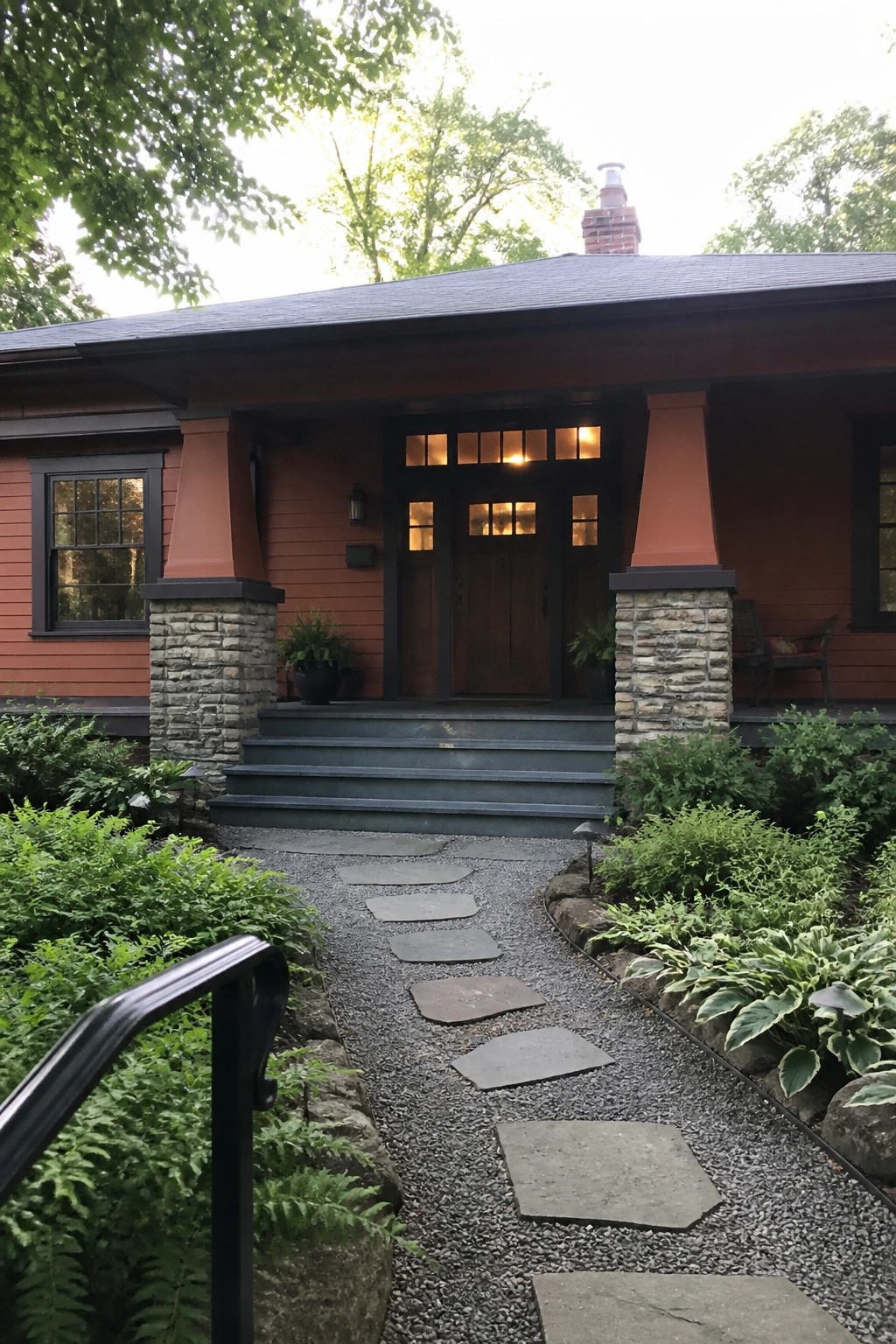 Red house with stone pillars, dark door, and gravel path amid greenery.