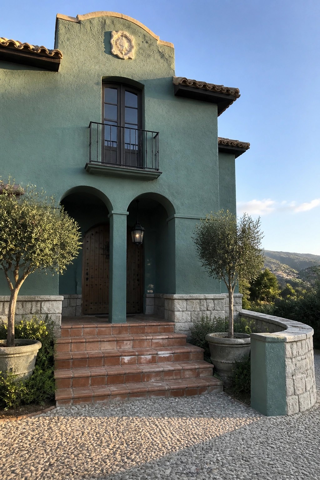 Teal stucco house with arched entry, wooden doors, balcony, terracotta steps, and potted trees.