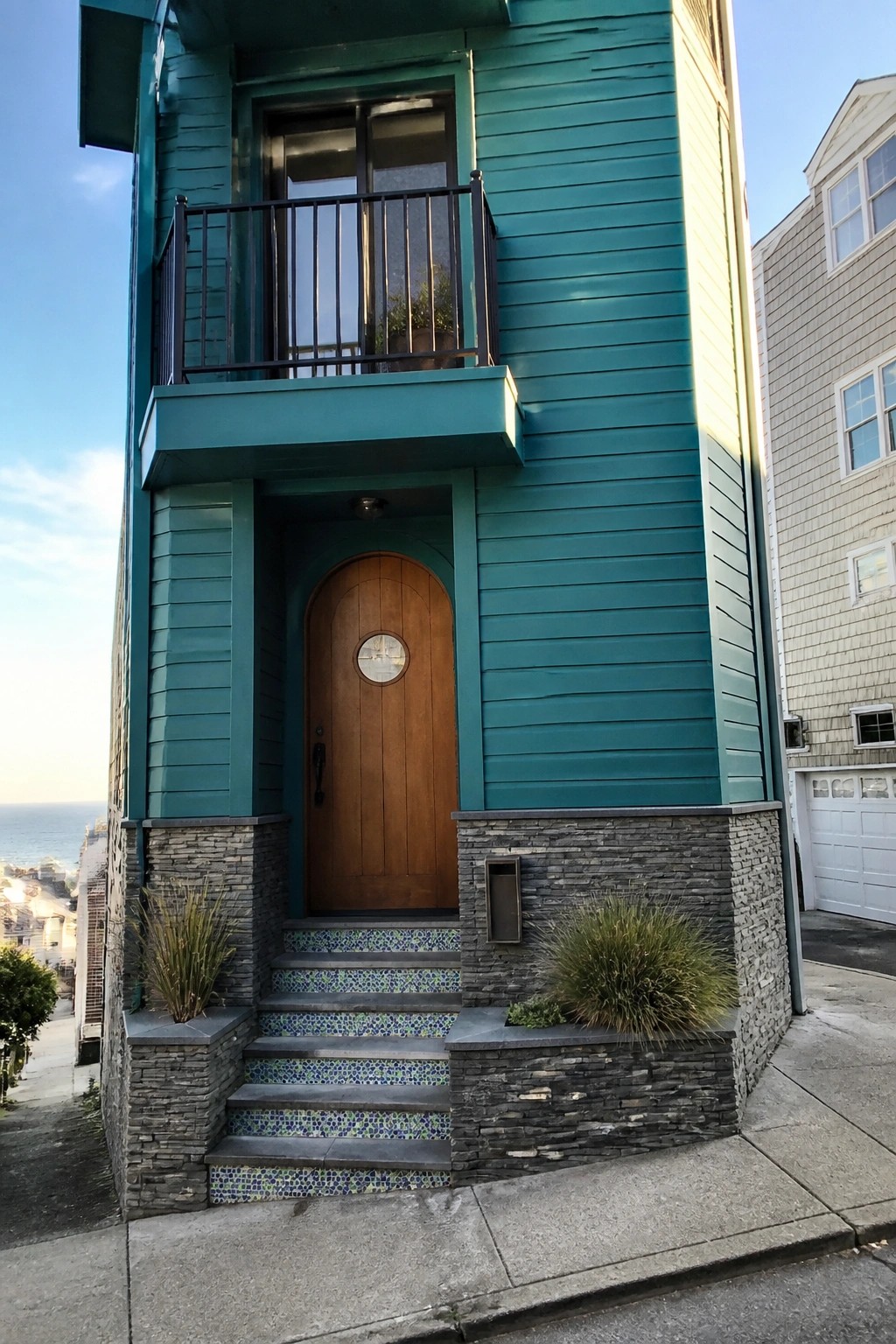 Teal house with arched wooden door, mosaic-tiled steps, balcony, and ocean view.