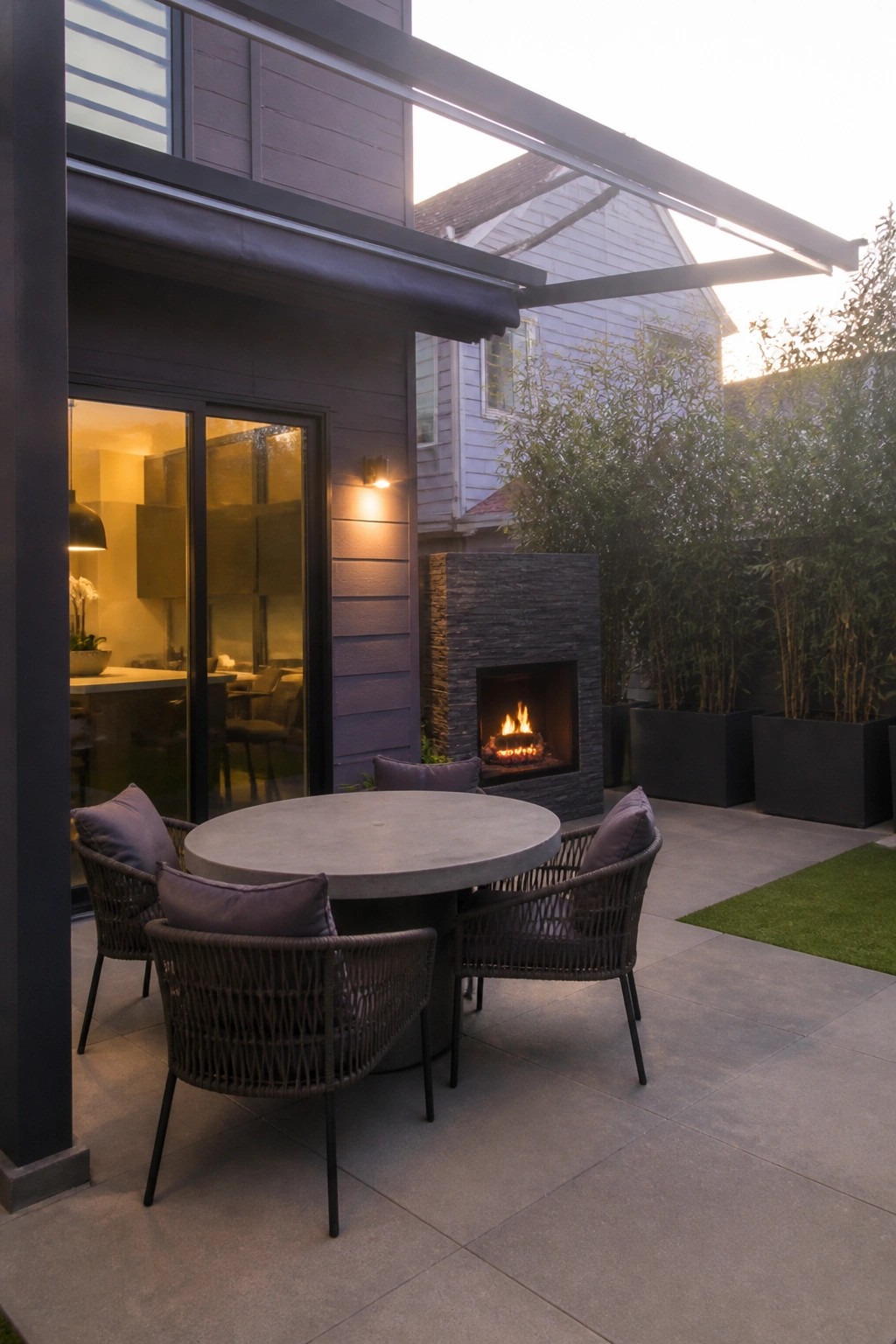 Outdoor patio with round concrete table, four woven chairs with cushions, and a stone fireplace set into the side of a house with dark siding.
