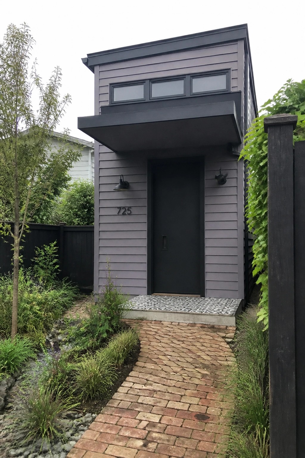 Modern narrow house with dark gray siding, black door, and brick pathway