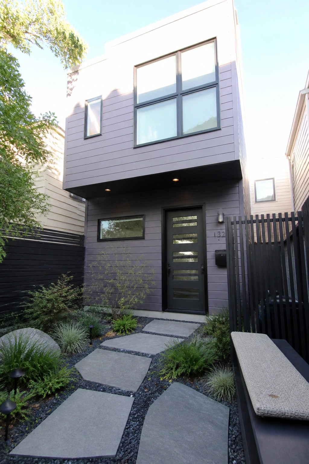 A narrow two-story house with purple-gray horizontal siding, large black-framed windows on the upper level, a black front door with horizontal slats, and a stone paver path through gravel and low plants.