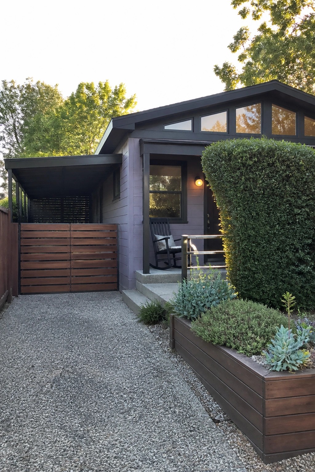 A single-story house with purple-gray siding, charcoal roof and trim, a covered entry porch, and a gravel driveway beside a wooden planter.