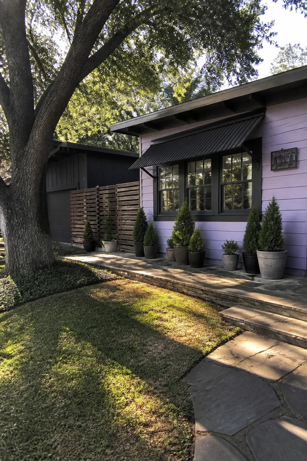 A light purple-gray house with black window frames, a black corrugated awning, and rows of potted evergreen shrubs along a stone path.