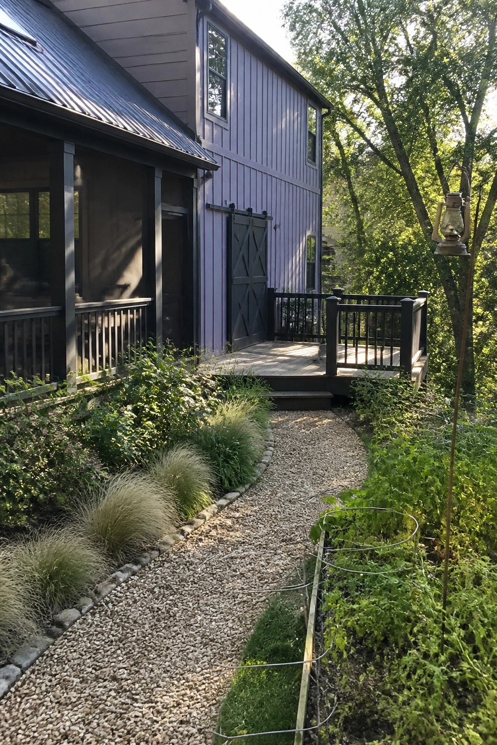 A two-story house with light purple-gray vertical siding, charcoal doors and trim, a wooden deck, and a gravel path bordered by plants.