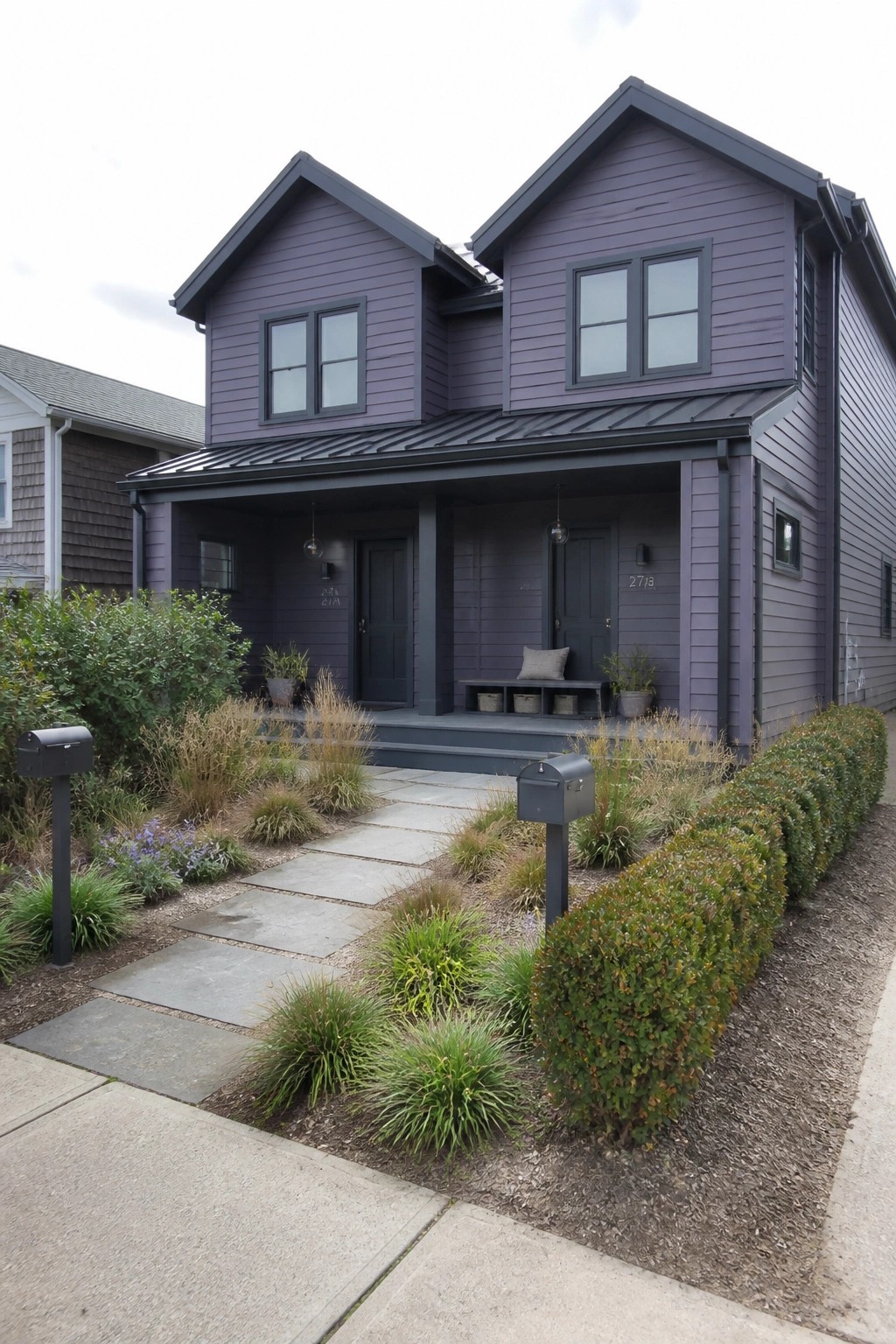 A two-story house with purple-gray siding, charcoal trim and metal roof, two dark front doors under a porch, and a stone path through the front garden.