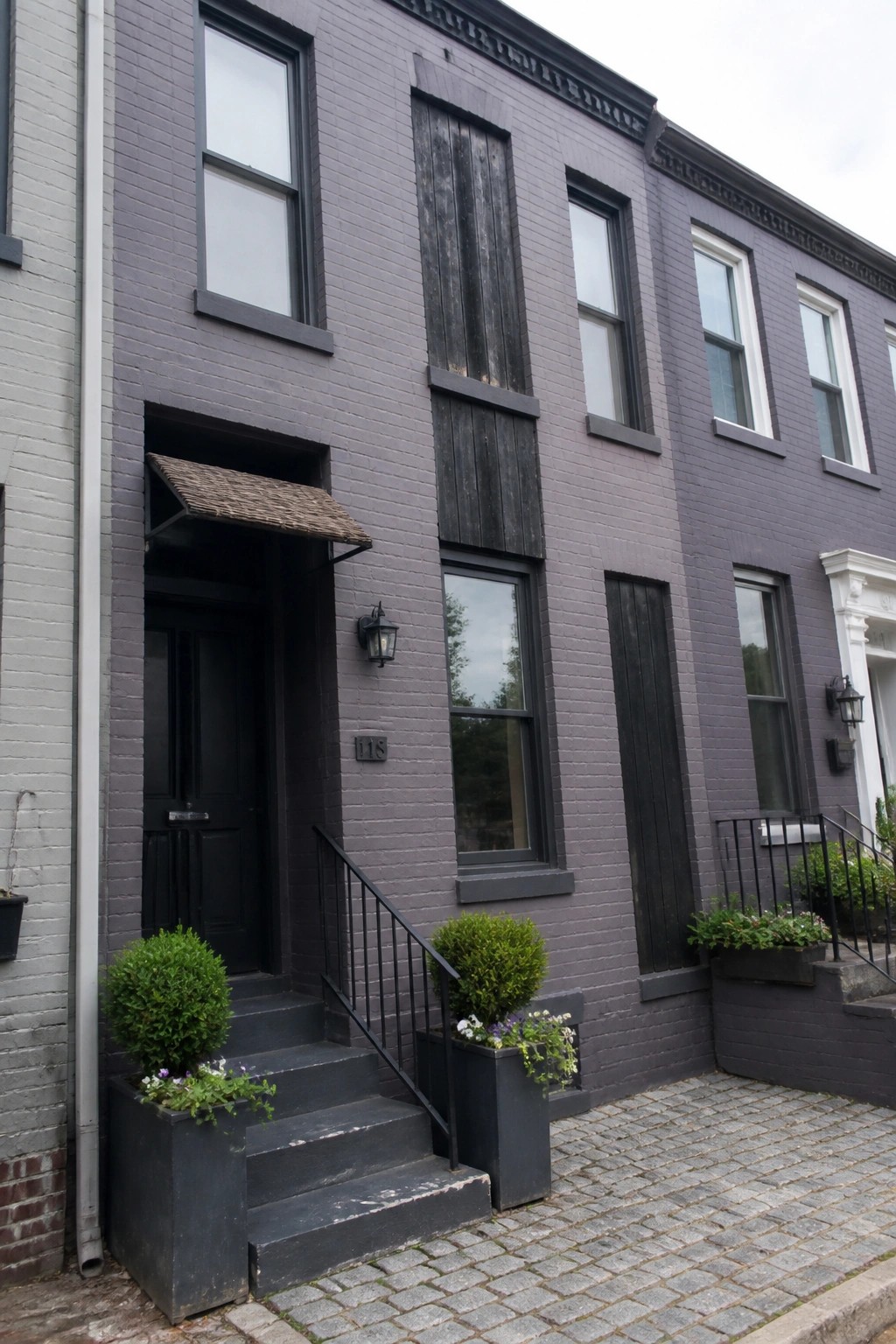 A purple gray brick row house exterior with black door, windows, and trim, black steps, and two large planters on a cobblestone sidewalk.
