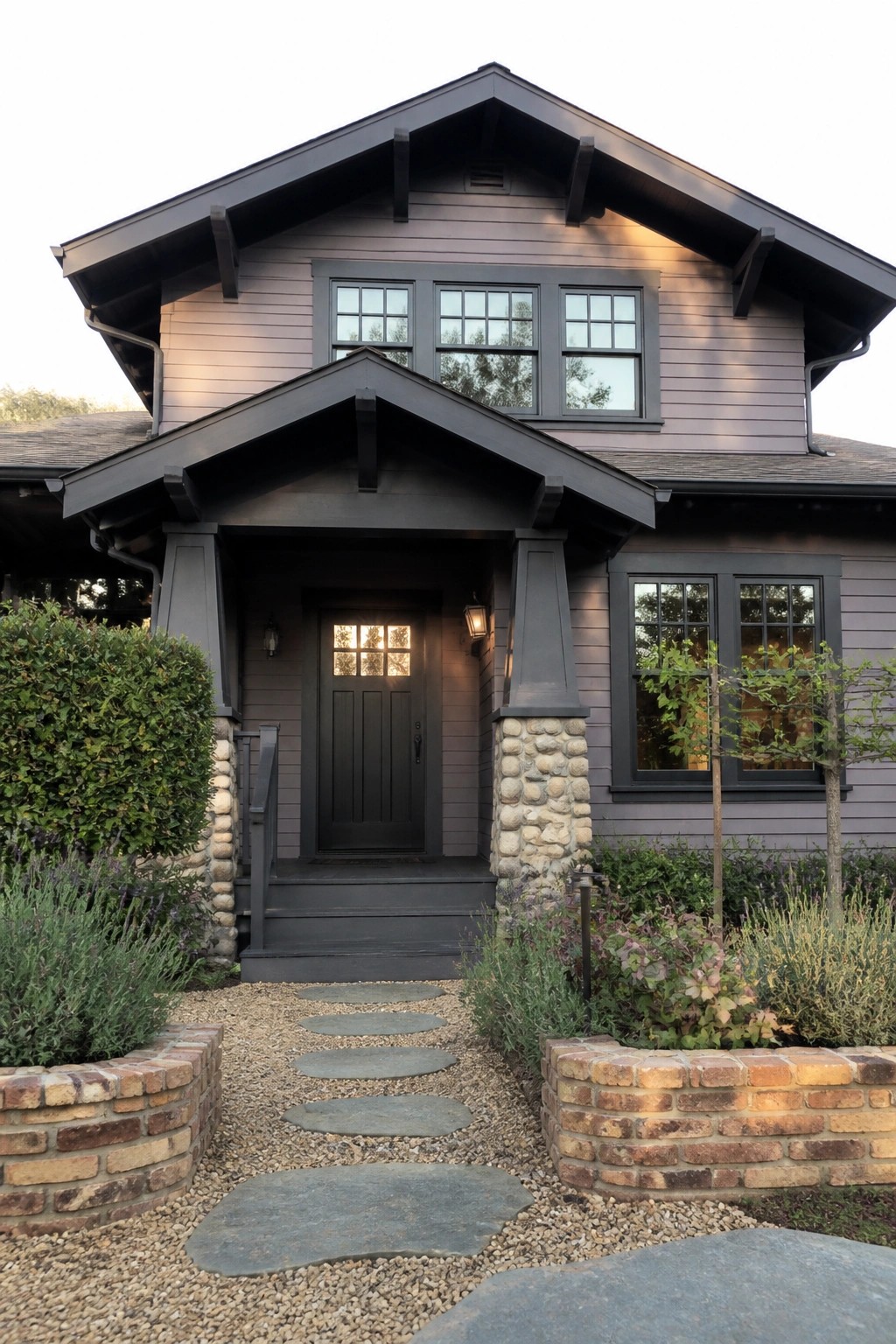Front view of a two-story house with purple-gray siding, charcoal trim, a dark paneled door, stone porch supports, and a stone pathway through gravel landscaping.