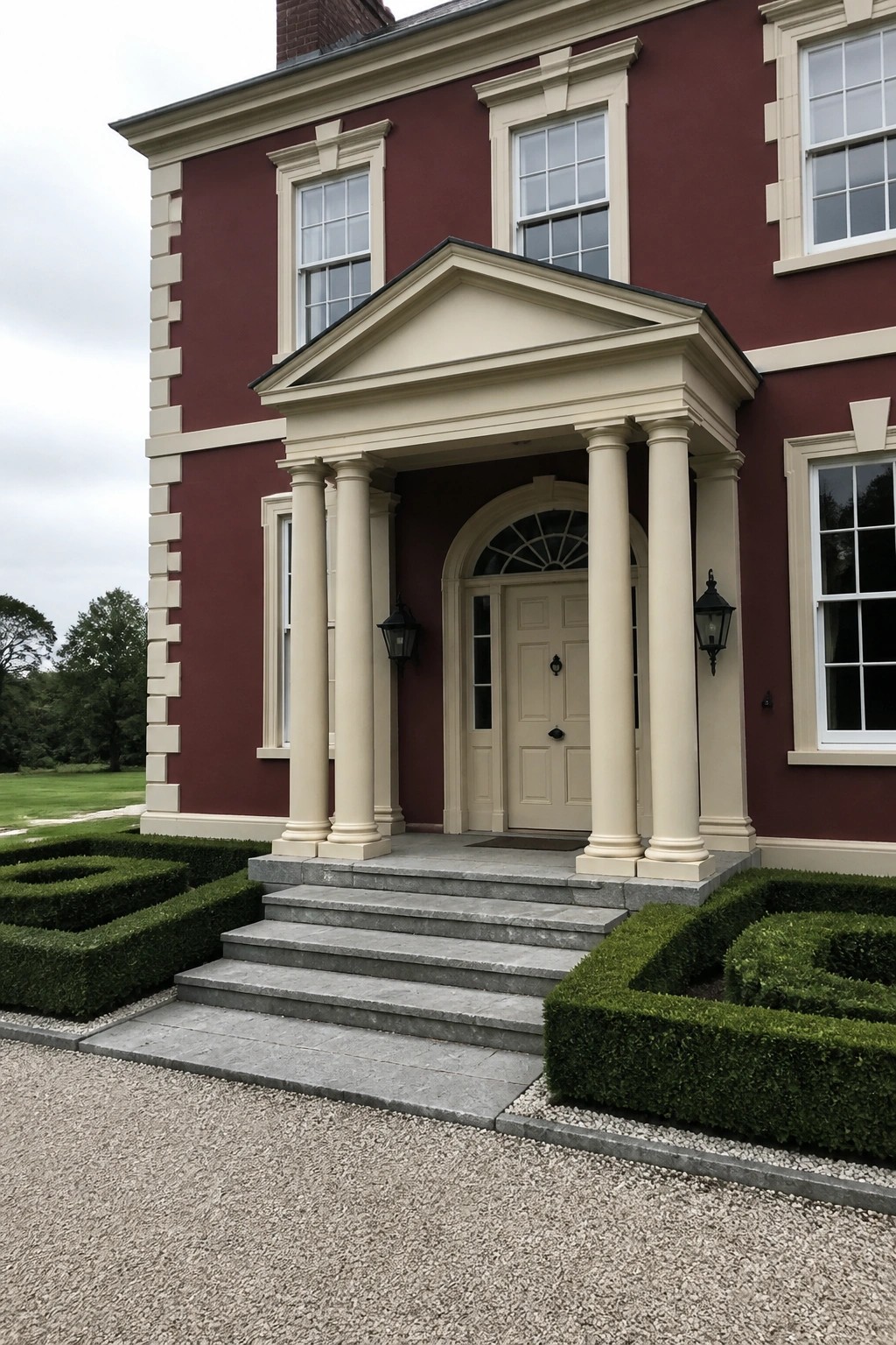 A two-story wine red house with cream trim shows a classical portico with four columns over the front door, stone steps, and boxwood hedges on either side.