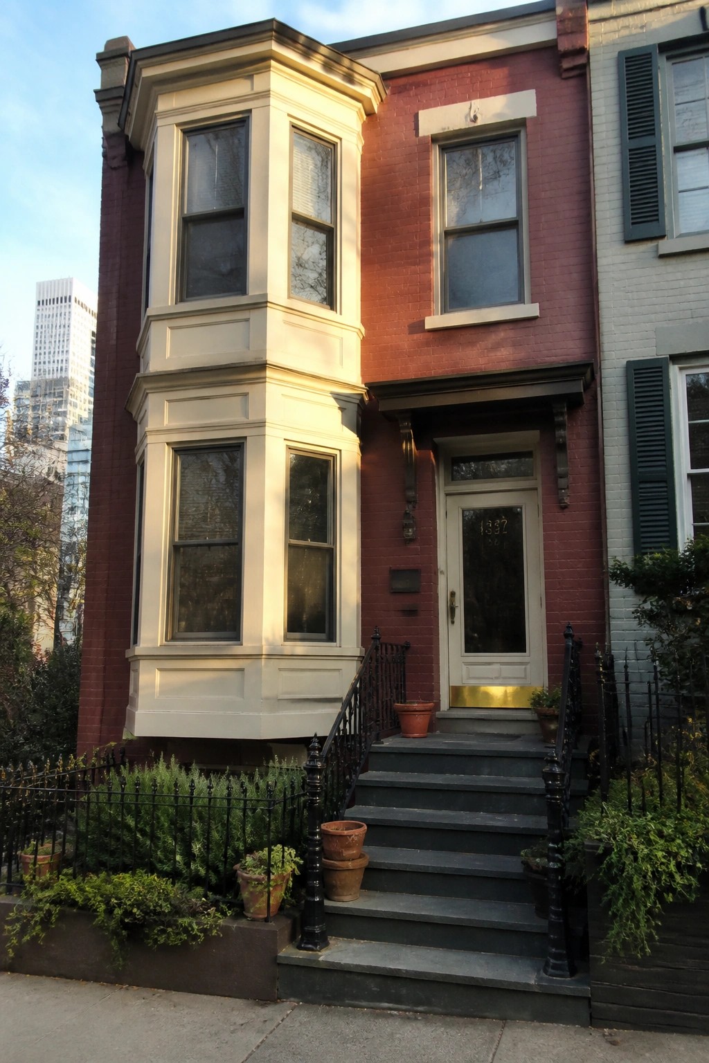 Red brick townhouse with white bay window, dark stairs, and potted plants.