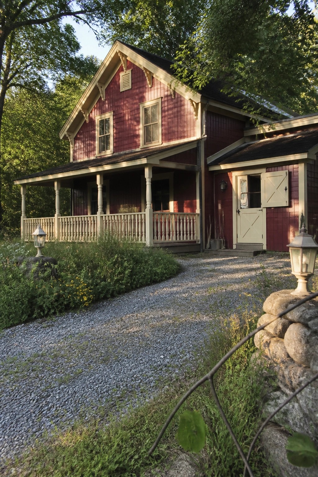 Red wooden farmhouse with white porch along gravel driveway amid trees and rocks