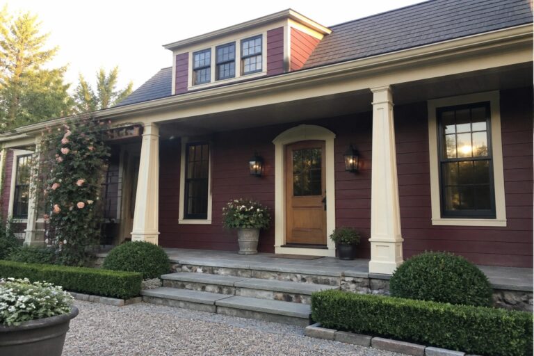 A two-story house with red siding, cream trim on the porch columns and window frames, a stone porch with steps, and a wooden front door.