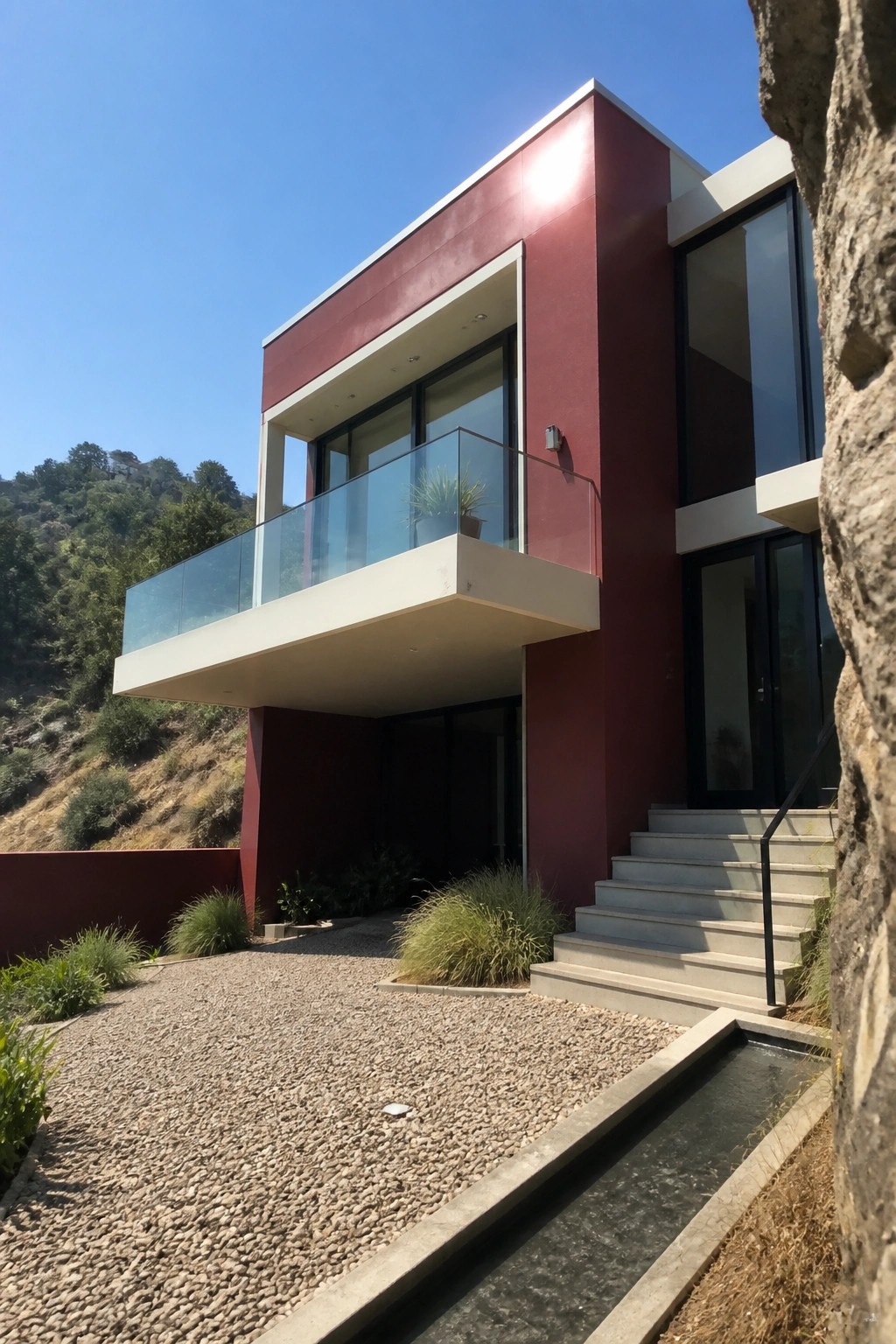 A modern two-story house with deep red exterior panels and cream trim, featuring a cantilevered balcony with glass railing and concrete stairs on a gravel driveway.
