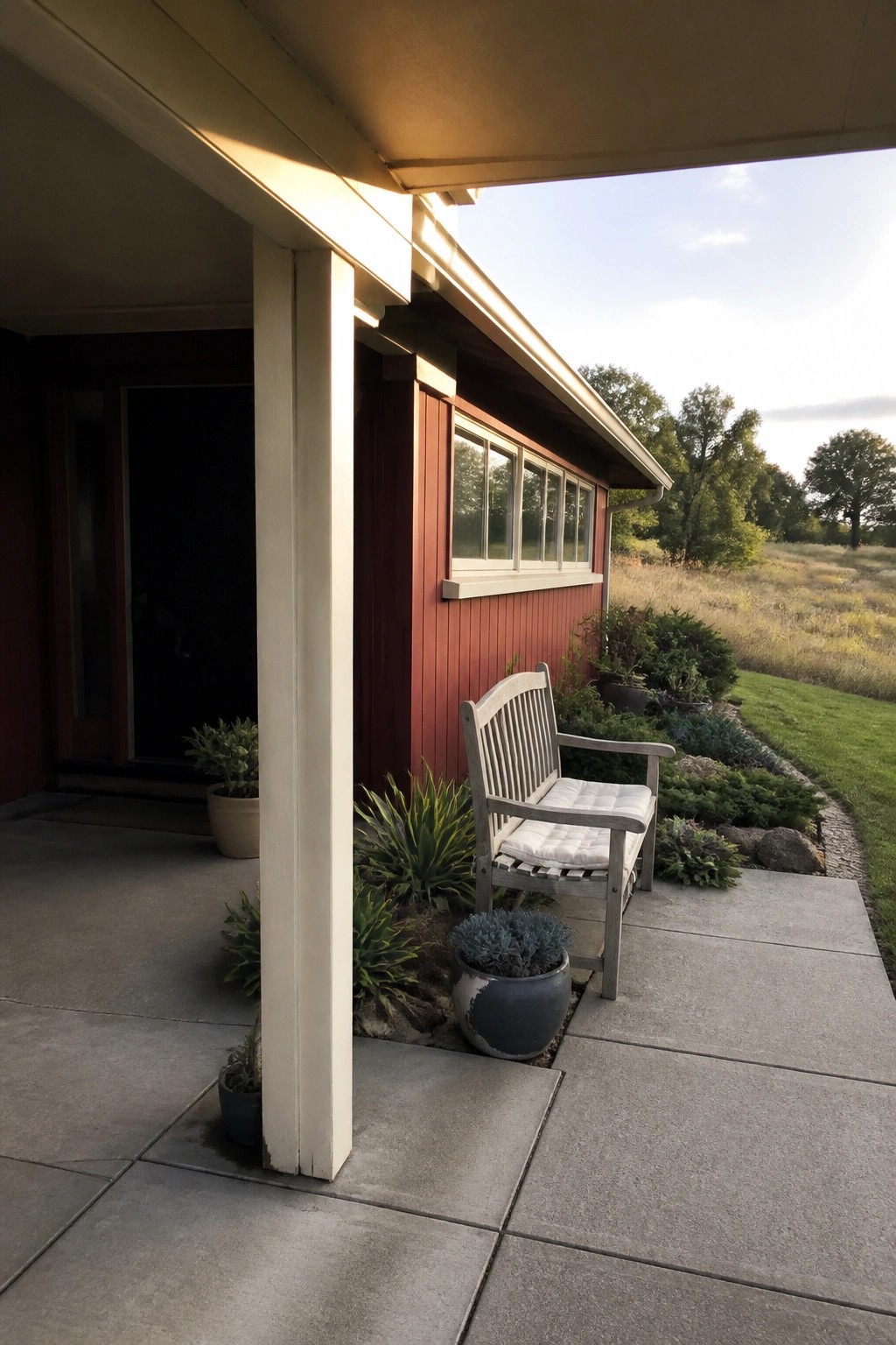 A red wooden house exterior with cream trim and pillars shows a covered porch area with a wooden bench and potted plants next to a grassy field.