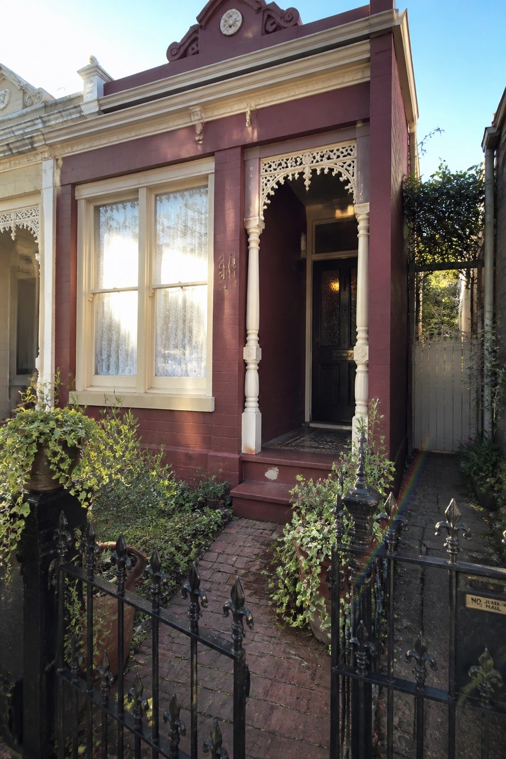 A wine-red painted brick house with cream trim around the windows and porch, a black front door, and a small front garden with potted plants behind a black iron fence.