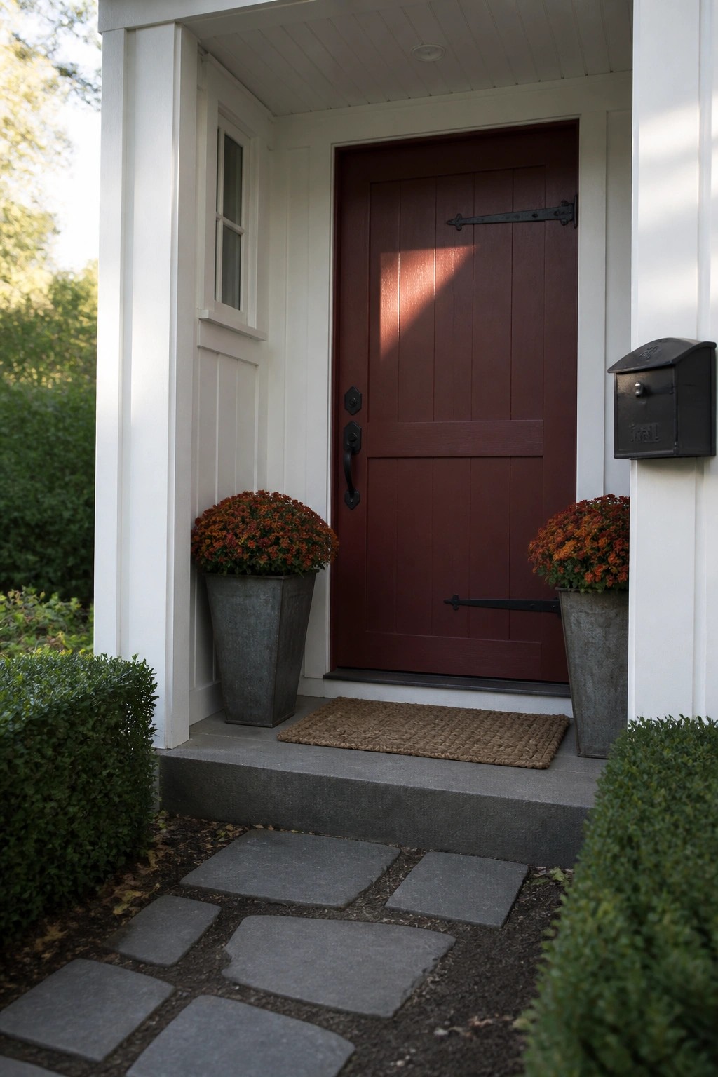 Red door on white porch flanked by orange mums in metal pots and stone path
