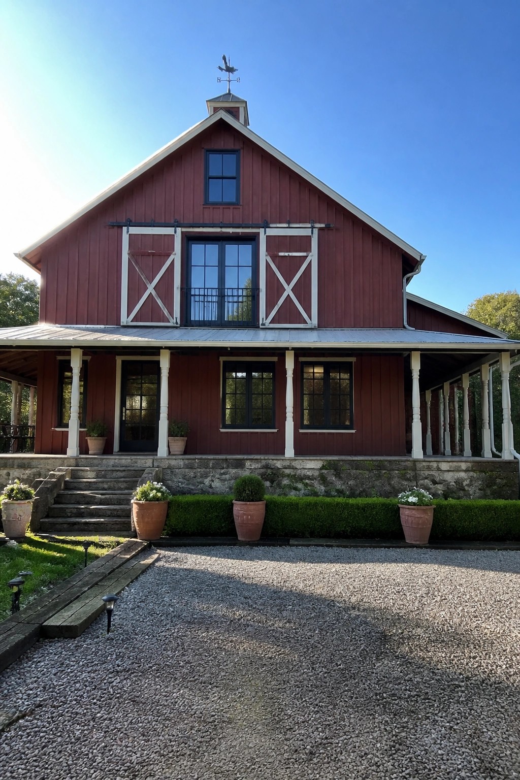 A two-story red barn-style house with white board-and-batten doors, black-framed windows, a metal roof, stone foundation, and a covered porch with white columns.