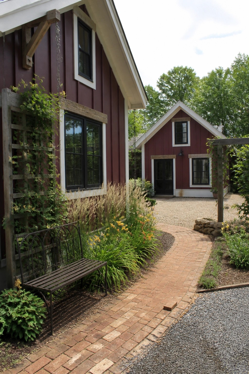 A burgundy red farmhouse exterior with white trim, black windows, a brick path, and garden beds along the side.