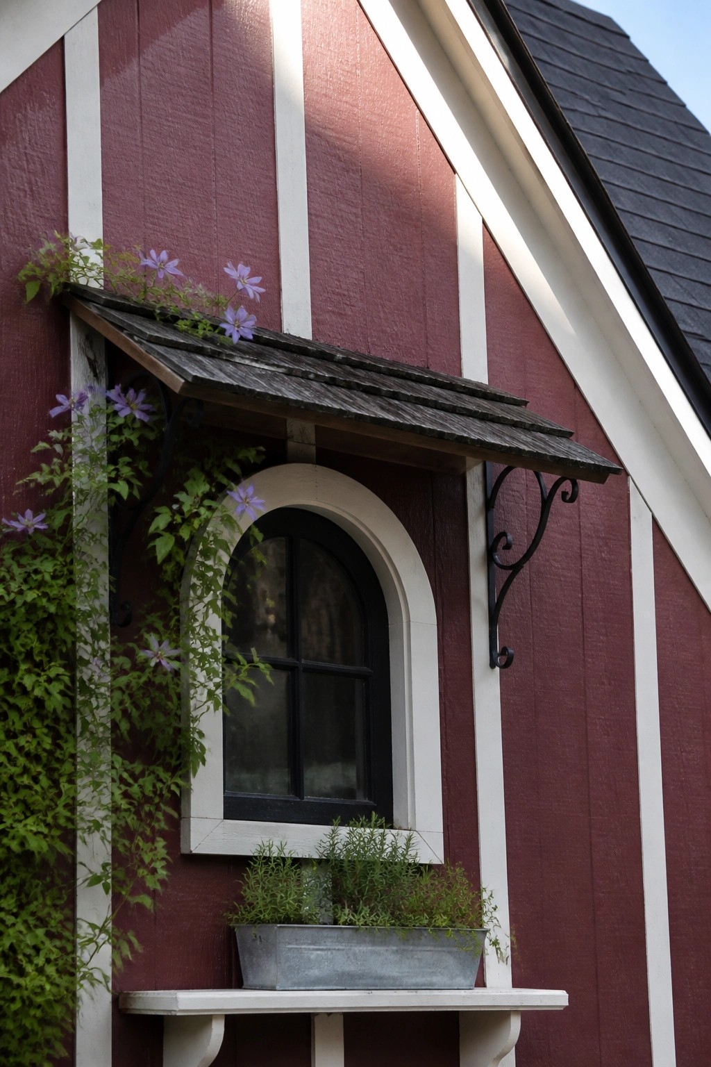 A close view of a burgundy wood-sided house wall with white vertical trim, an arched black window under a small shingled awning, and a metal planter on a white ledge.