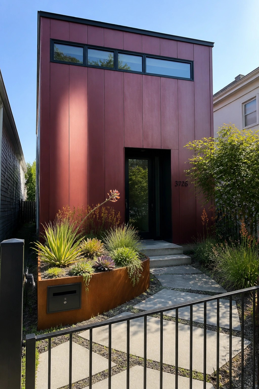 A narrow two-story modern house with matte burgundy metal siding, black-framed windows, and a black front door, shown with a concrete path and rusted steel planter bed in the front yard.