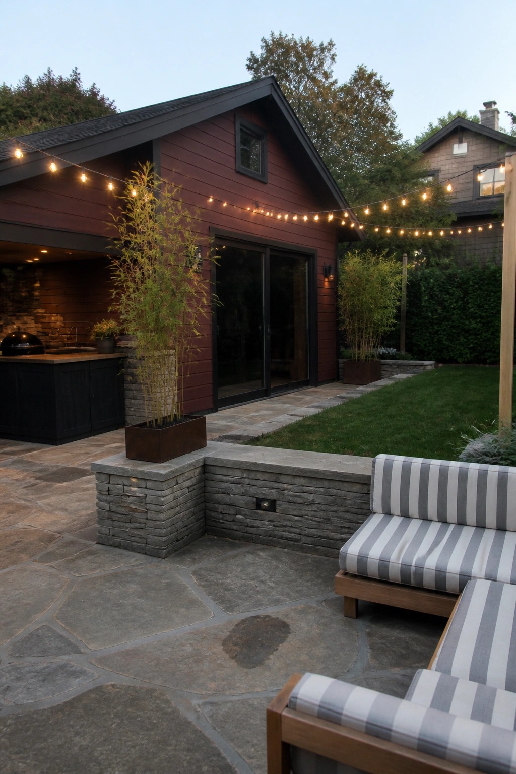 A burgundy house exterior with black trim, sliding glass doors, and an attached stone patio with striped seating and string lights.