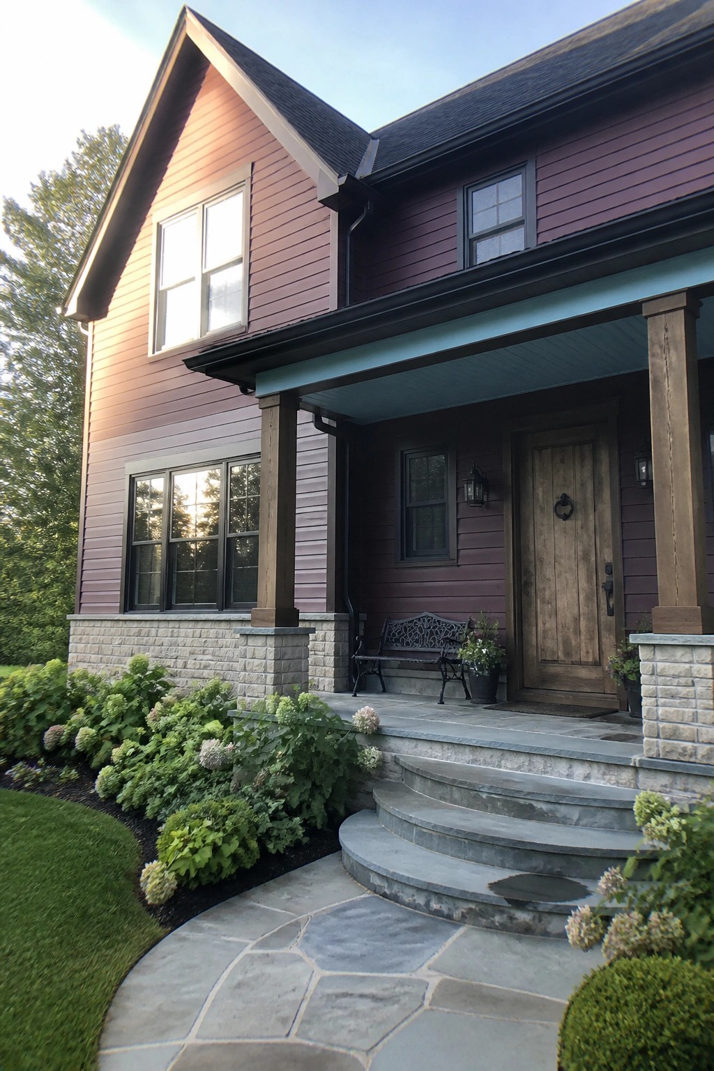 A two-story house with deep maroon lap siding, a stone foundation, a wooden front door under a covered porch, and stone steps leading up from a curved stone walkway.