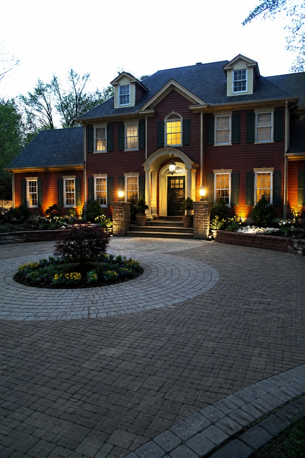 Red two-story house with arched entry, lit windows, and circular brick driveway at dusk